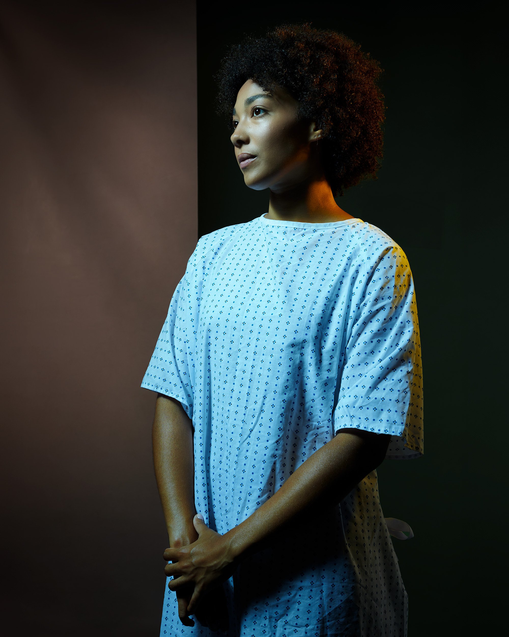 Professional commercial portrait of a woman with natural curly hair wearing a hospital gown, standing with hands clasped in front of her, in a photo studio sets as an hospital.