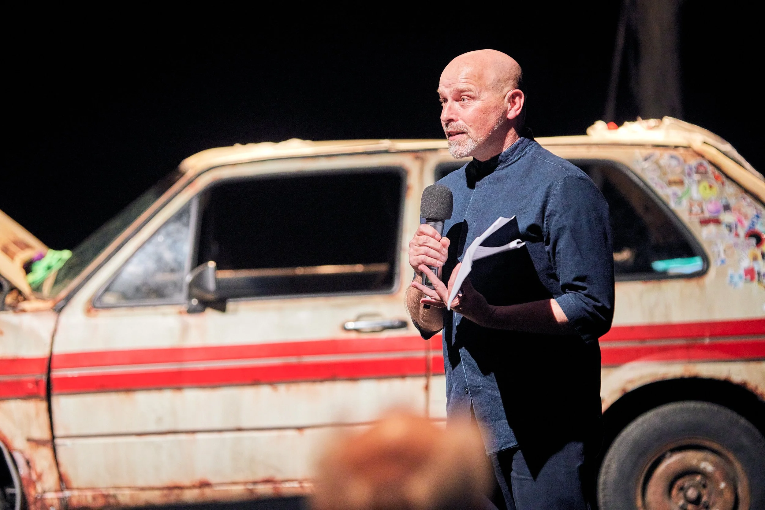 A man holding a microphone and notes speaking on stage with an old, rusty car covered in stickers behind him.