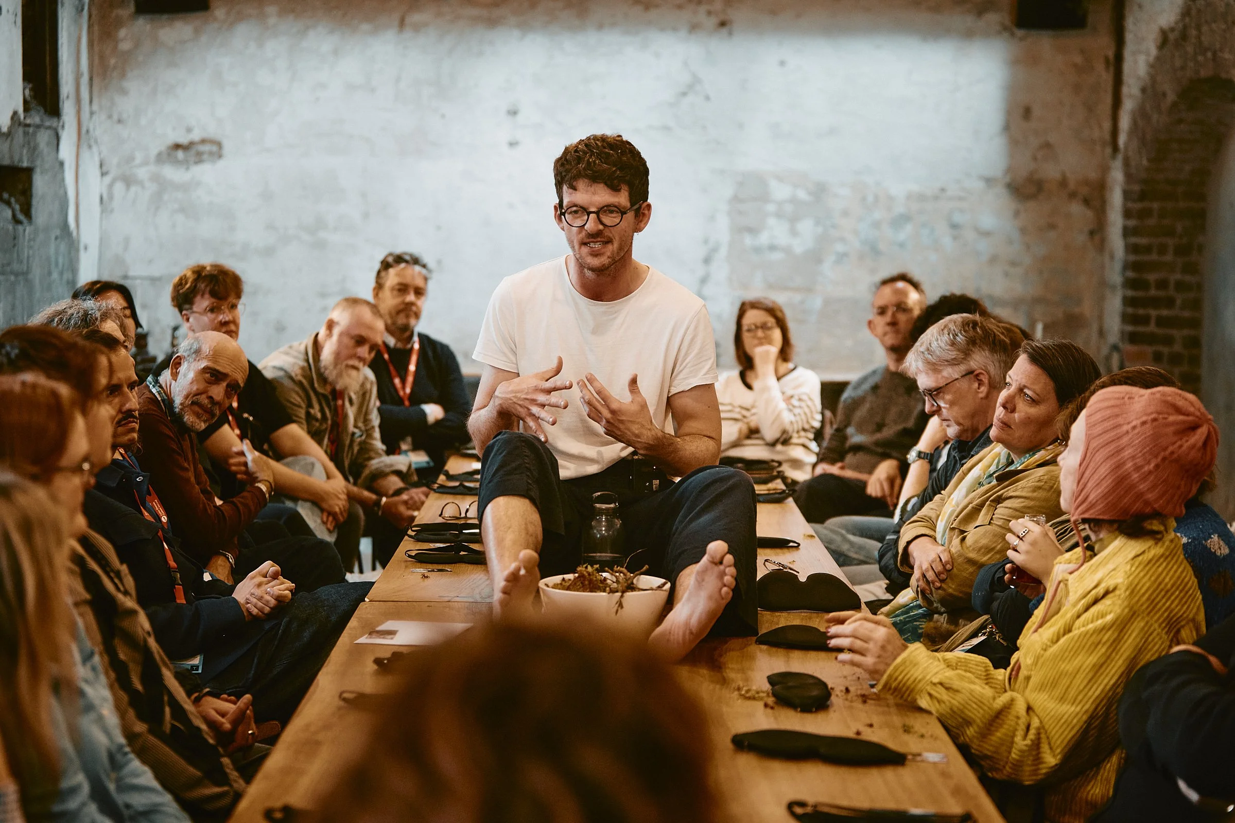 A man with glasses and a white t-shirt is sitting cross-legged on a large wooden table, speaking to a group of people in a meeting or workshop setting. The group is seated around the table, listening attentively. The room has exposed brick and concre