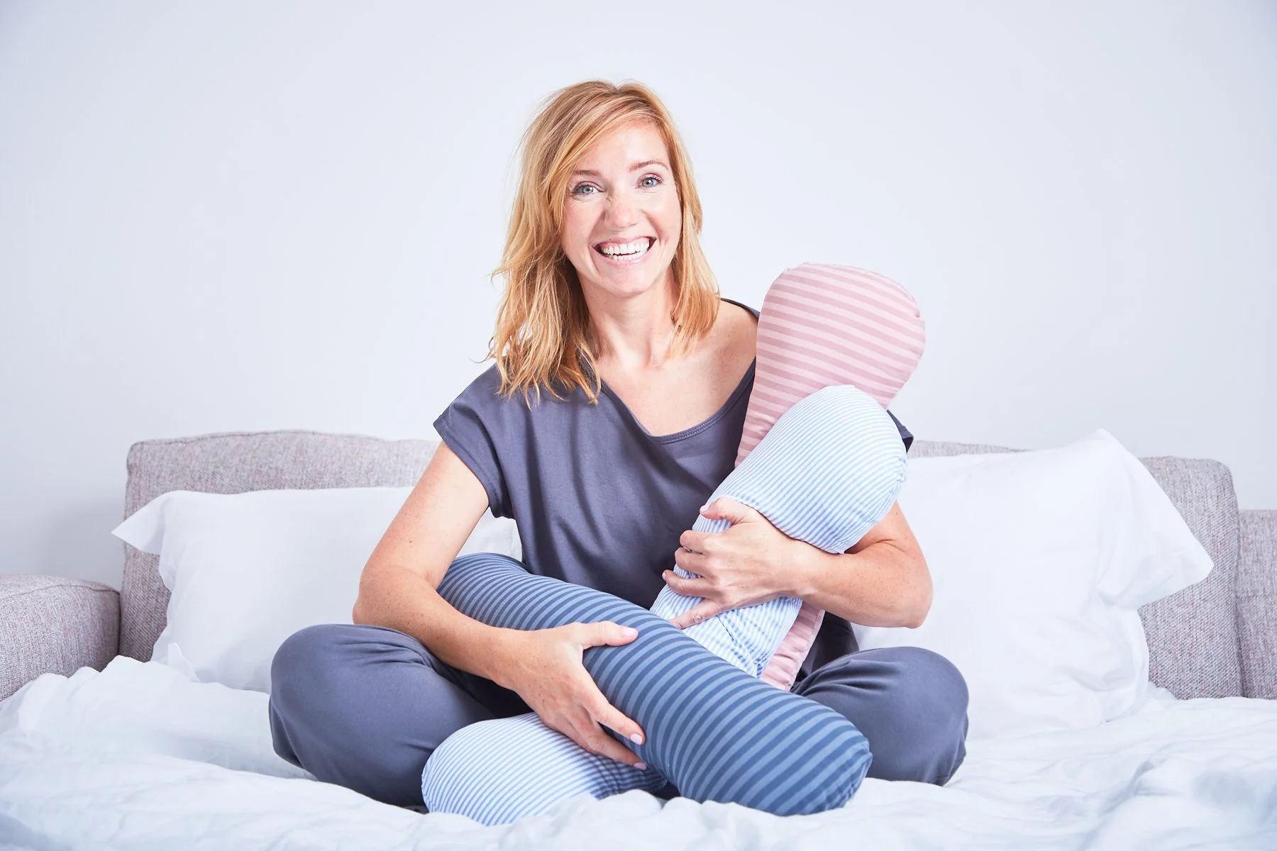 A woman with red hair sitting on a bed, smiling, holding two striped pillows, one pink and one blue.