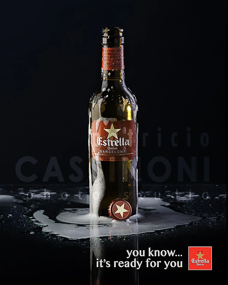 Advertising profession product photo taken in studio of a bottle of Estrella beer with foam around its base, set against a dark background with foam and reflections on the surface.