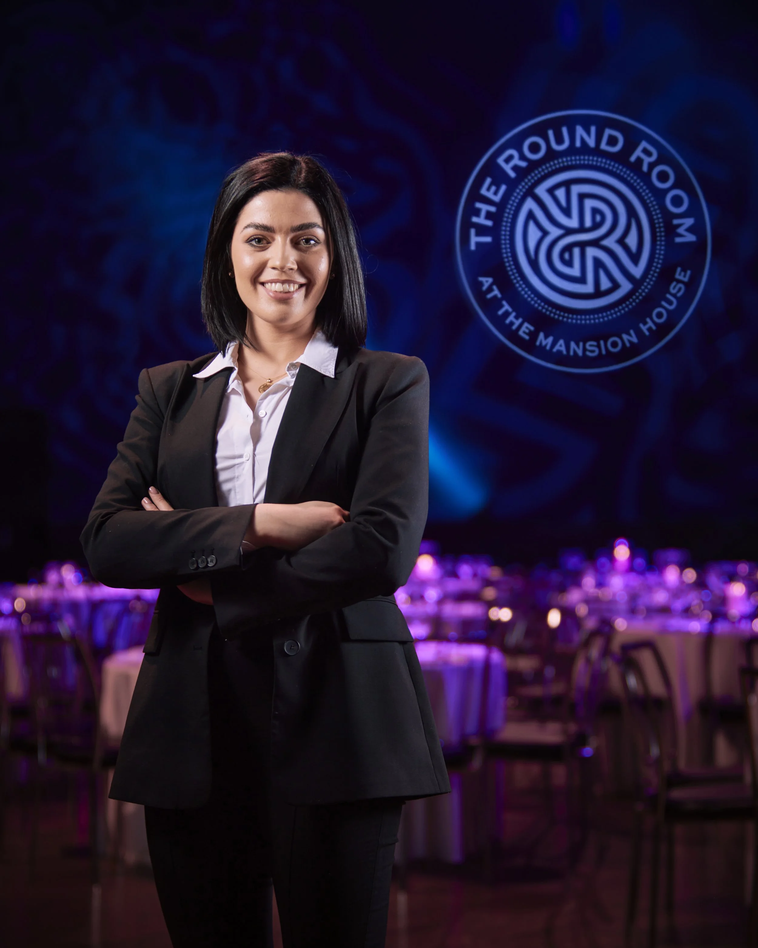 Corporate portrait photography of a woman in location with black hair in a business suit standing with arms crossed in a dimly lit event space, with purple and blue lighting.