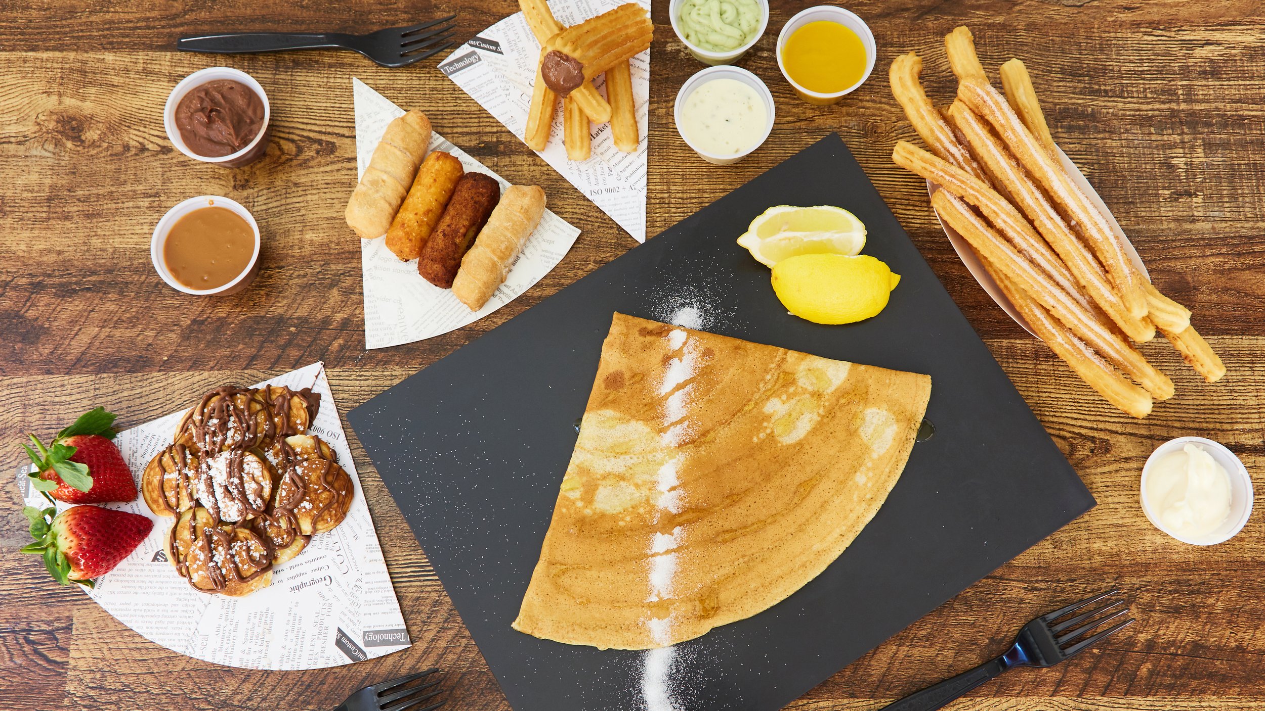 Advertising commercial professional photo of a variety of desserts and fried foods on a wooden table, including a large crepe with lemon slices, churros, strawberries with chocolate drizzle, and other fried items with dipping sauces.
