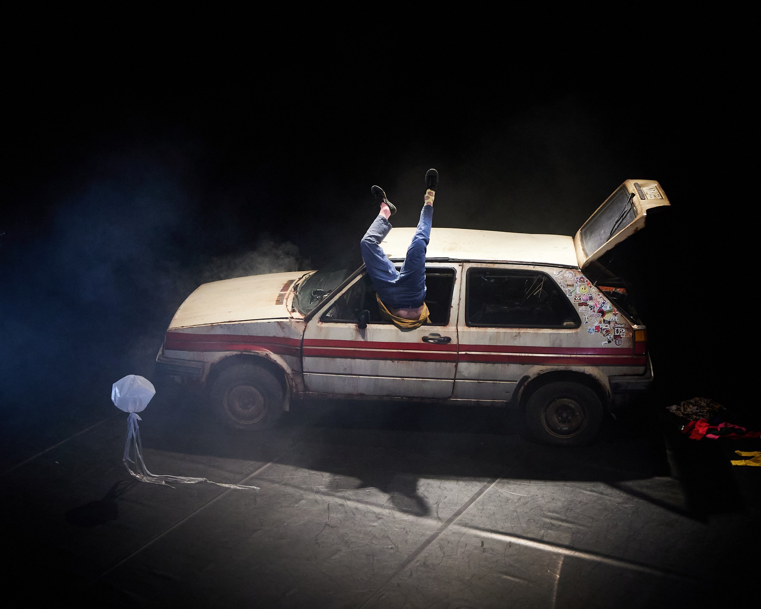 A person doing a handstand in the open sunroof of an old, dirty car decorated with stickers, parked on a dark stage with a white lantern nearby.