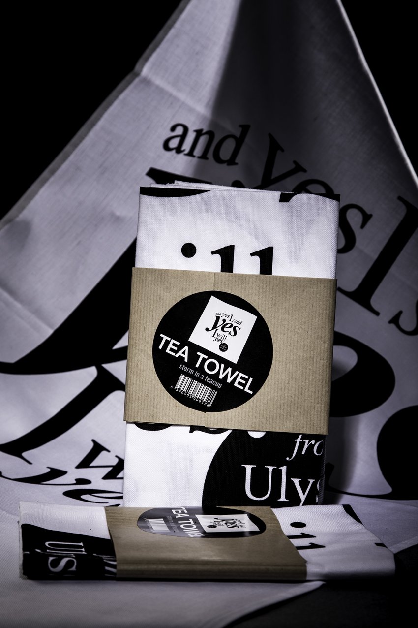 High end advertising profession product photo taken in studio of a tea towel with black and white text wrapped in a brown paper band, resting on a printed shopping bag and a folded towel display in the background.
