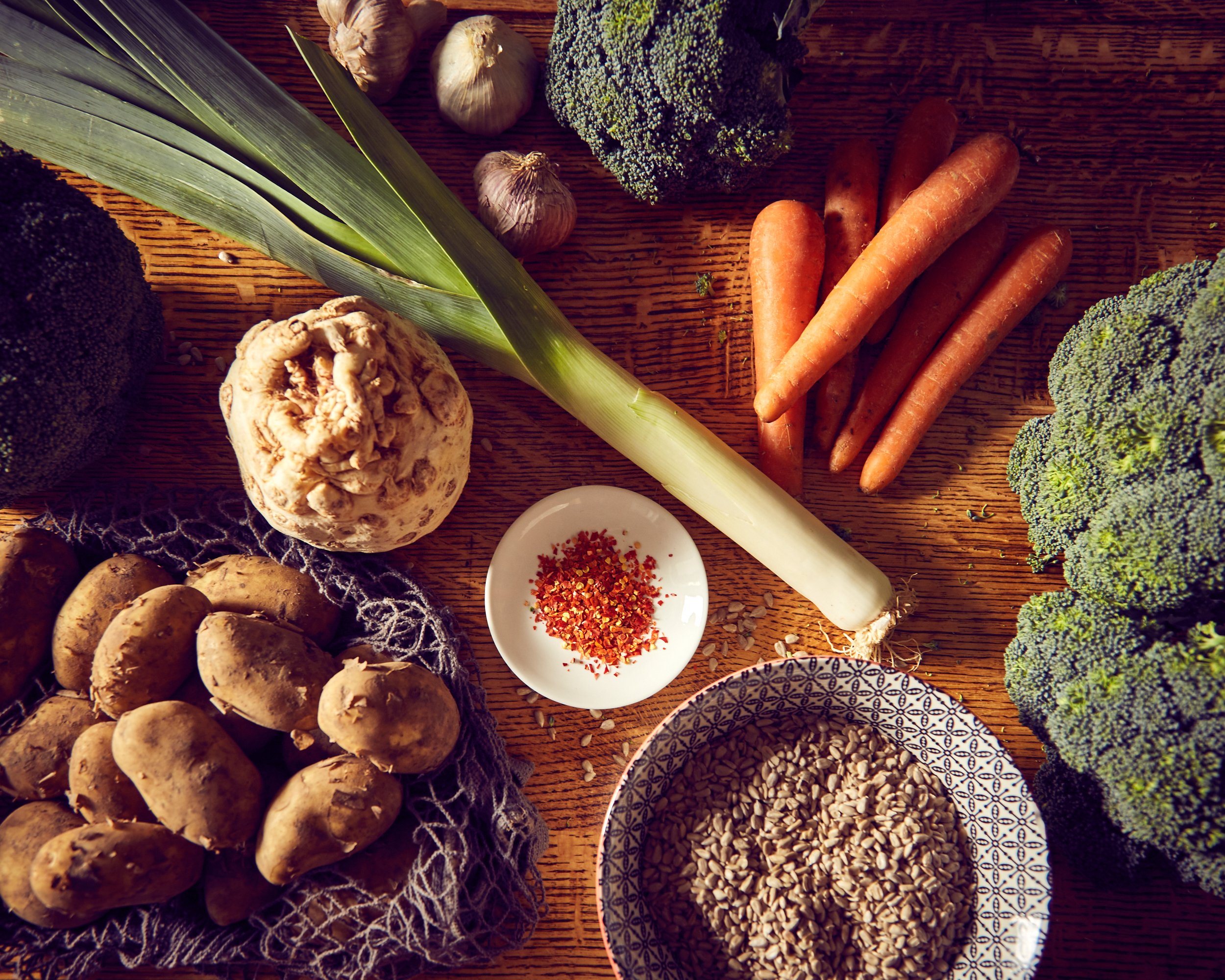 Fresh vegetables including carrots, broccoli, garlic, ginger, potatoes, leek, sunflower seeds, and red pepper flakes on a wooden surface.