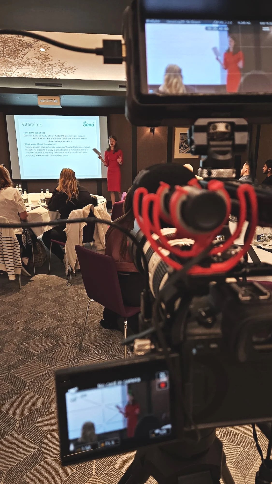 A woman in a red dress is giving a presentation in a conference room, with a large screen displaying text about Vitamin E behind her. The photo is taken from behind the camera, which is recording the event.