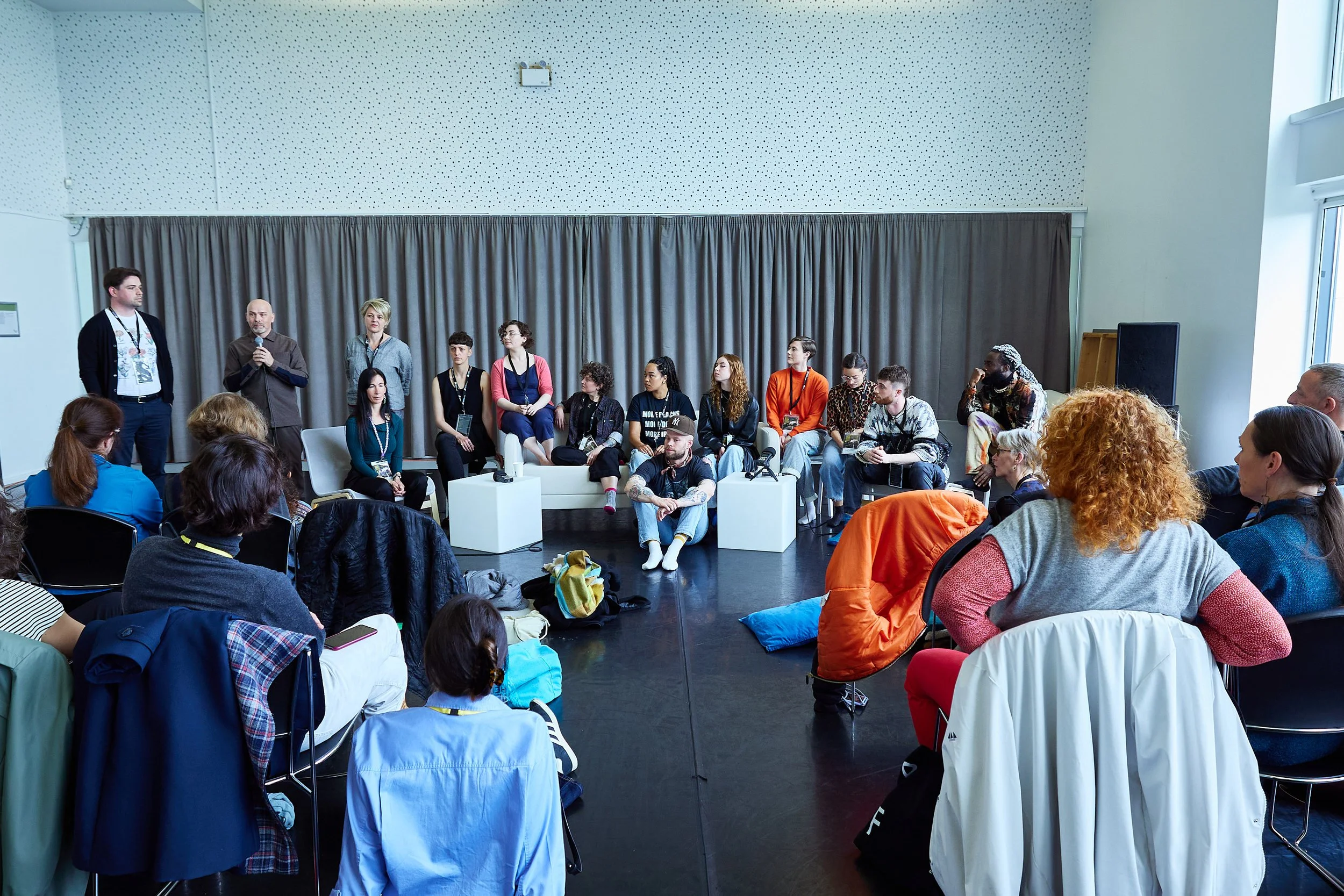 A panel discussion at a conference with diverse speakers and an audience seated in a room with large windows and curtains.