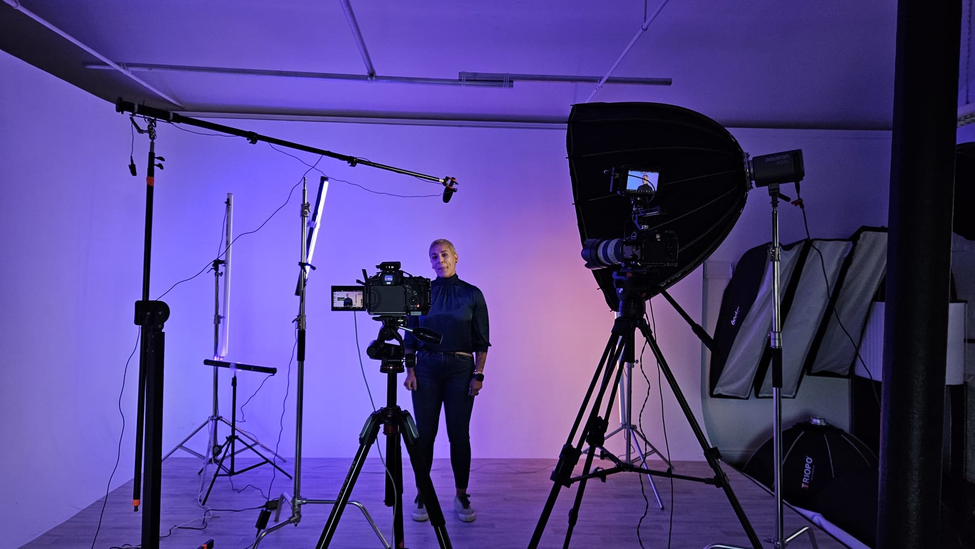 A woman stands in a professional photography or video studio surrounded by cameras, lights, and equipment, with a plain wall background and purple lighting.