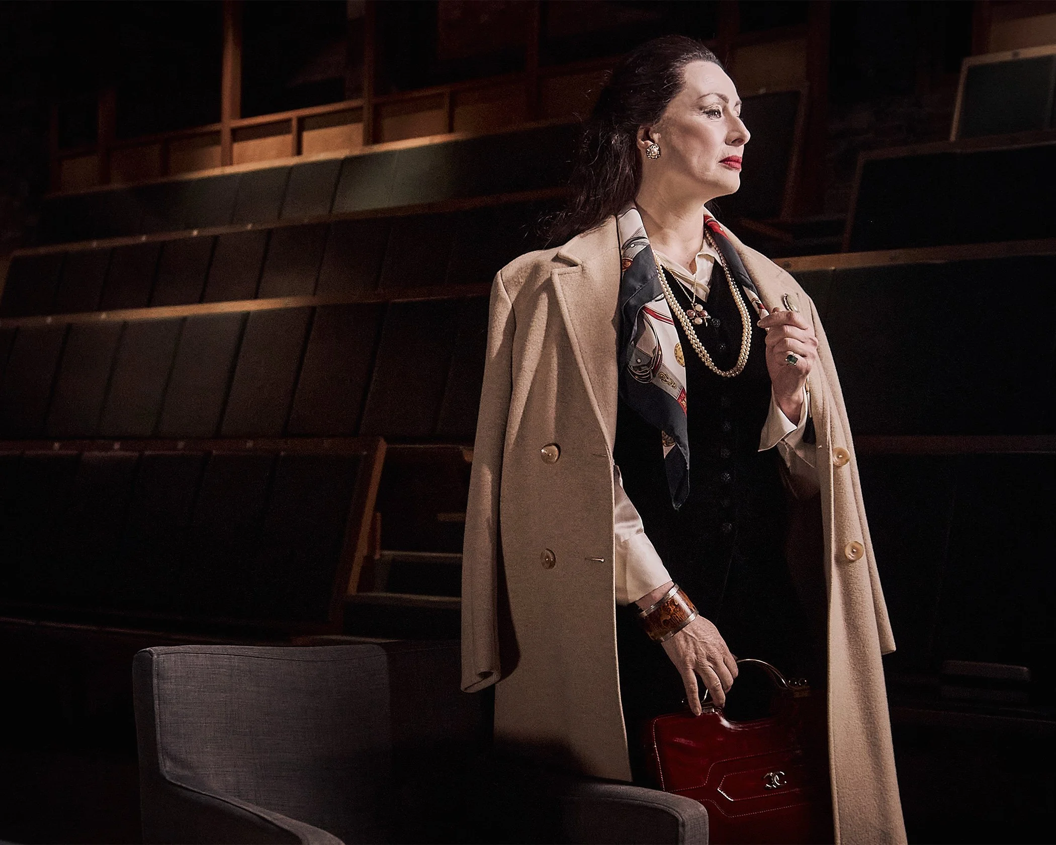 Advertising professional photo of an older woman impersonating Maria Calas, standing in an empty auditorium with wooden seats and a dark background, shooting in location with professional photography lighting.