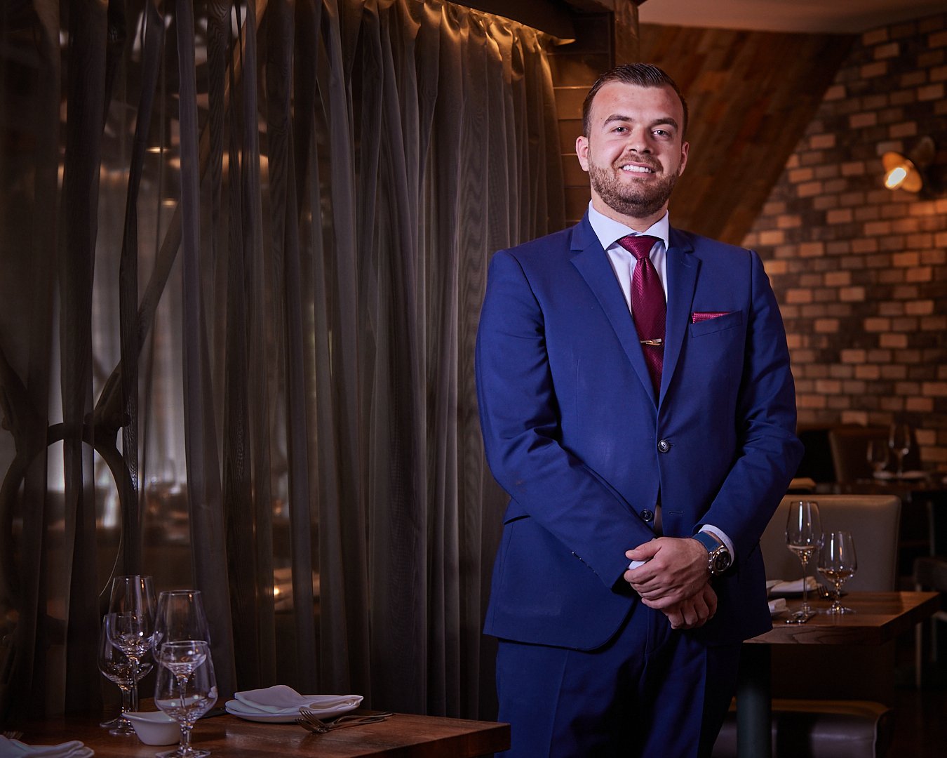 A man in a blue suit with a white shirt and burgundy tie standing inside a restaurant, smiling with arms crossed, with tables set with wine glasses, plates, and napkins in the background.