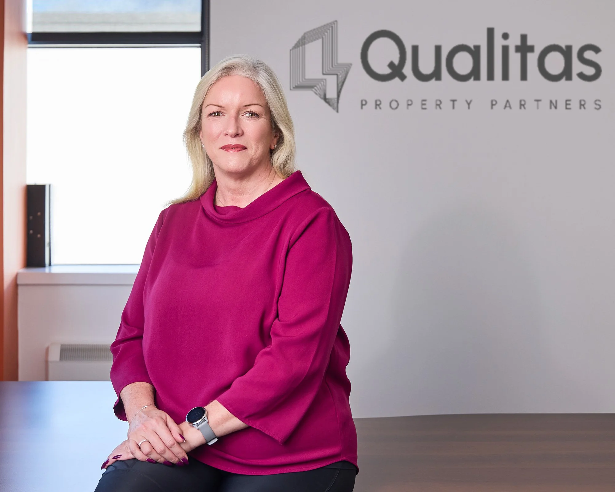 Professional corporate portrait, corporate headshot of a middle-aged woman with blonde hair in an office with a sign that reads 'Qualitas Property Partners' in the background, in Dublin.