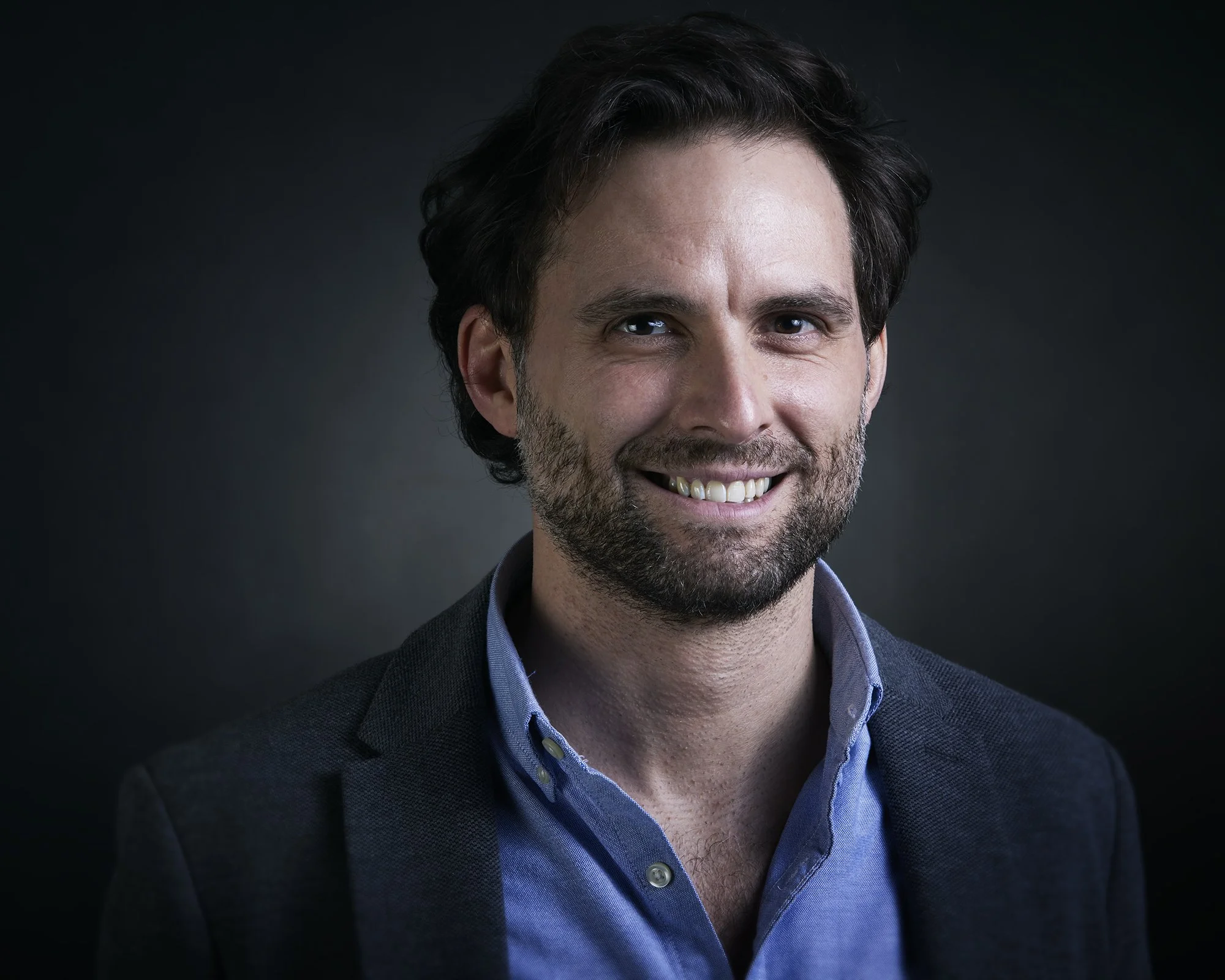A corporate headshot of a smiling professional man with dark, wavy hair and a beard, wearing a dark blazer over a blue collared shirt, against a dark background in a photo studio in a high quality lighted photo.