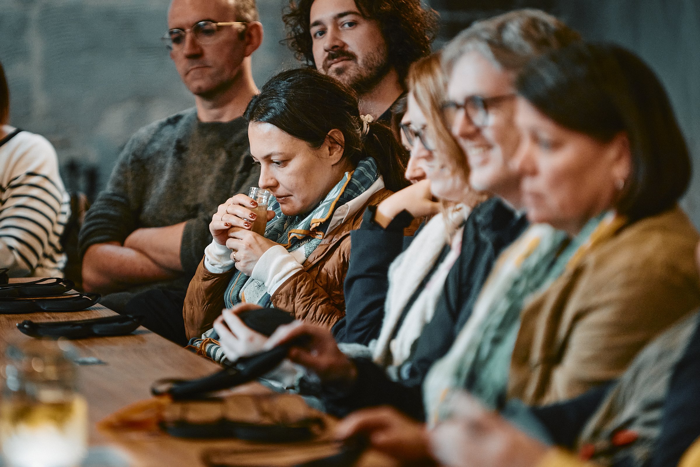 A group of people seated at a wooden table, some using smartphones and one woman drinking from a glass, in a dimly lit indoor setting.
