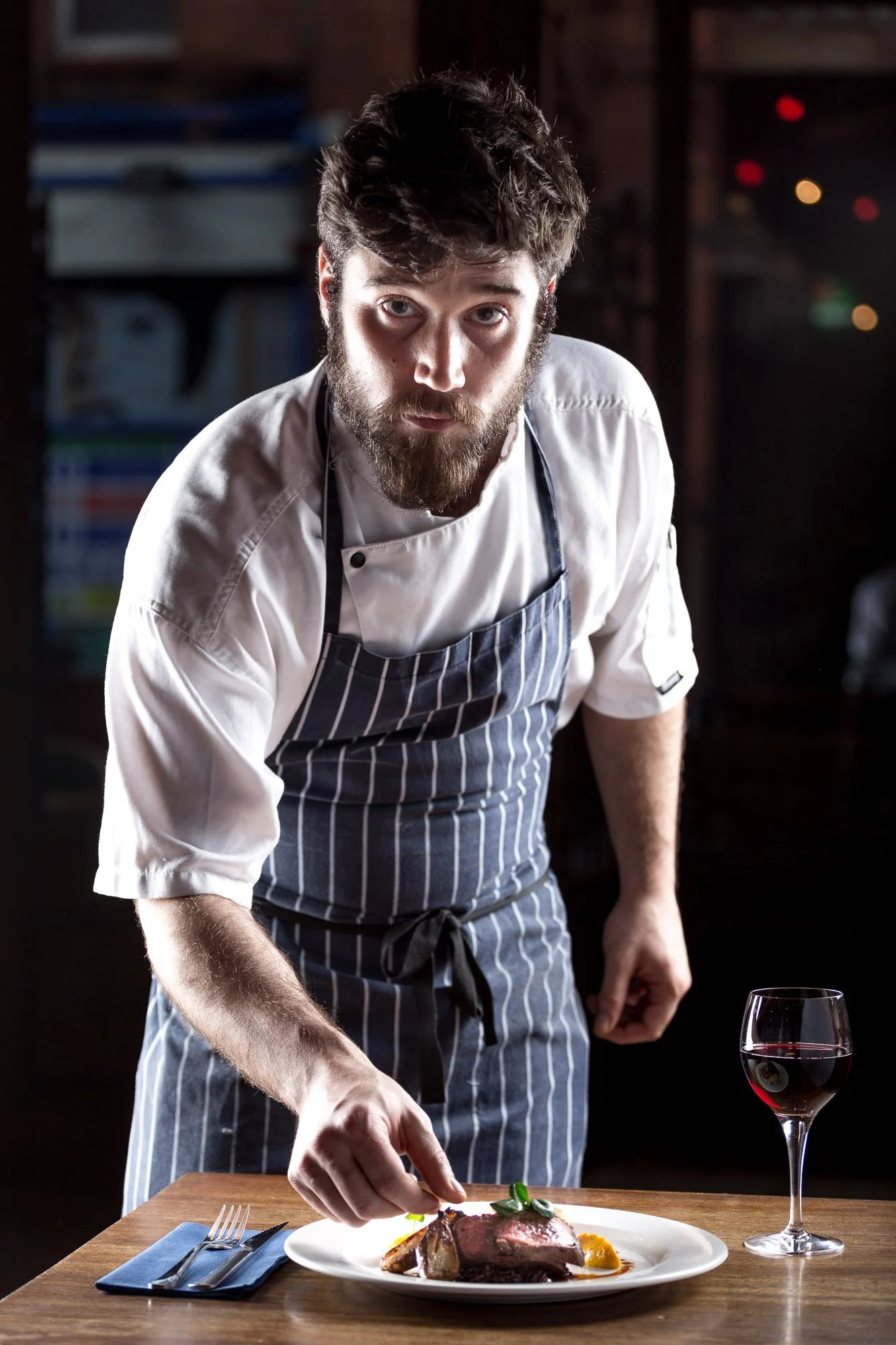 Advertising image of a chef carefully garnishes a plate of cooked meat with vegetables, with a glass of red wine on a wooden table, shooting in location with professional  photography lighting setup.