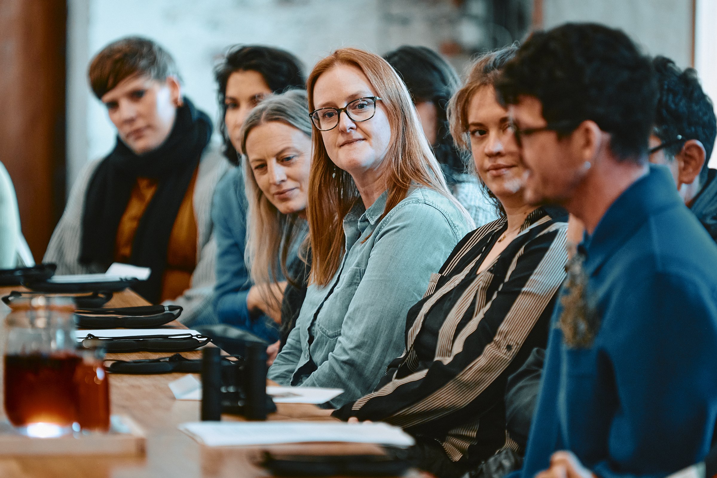 Group of people sitting at a conference table, attentively listening; some looking towards the camera, others focused on the speaker.