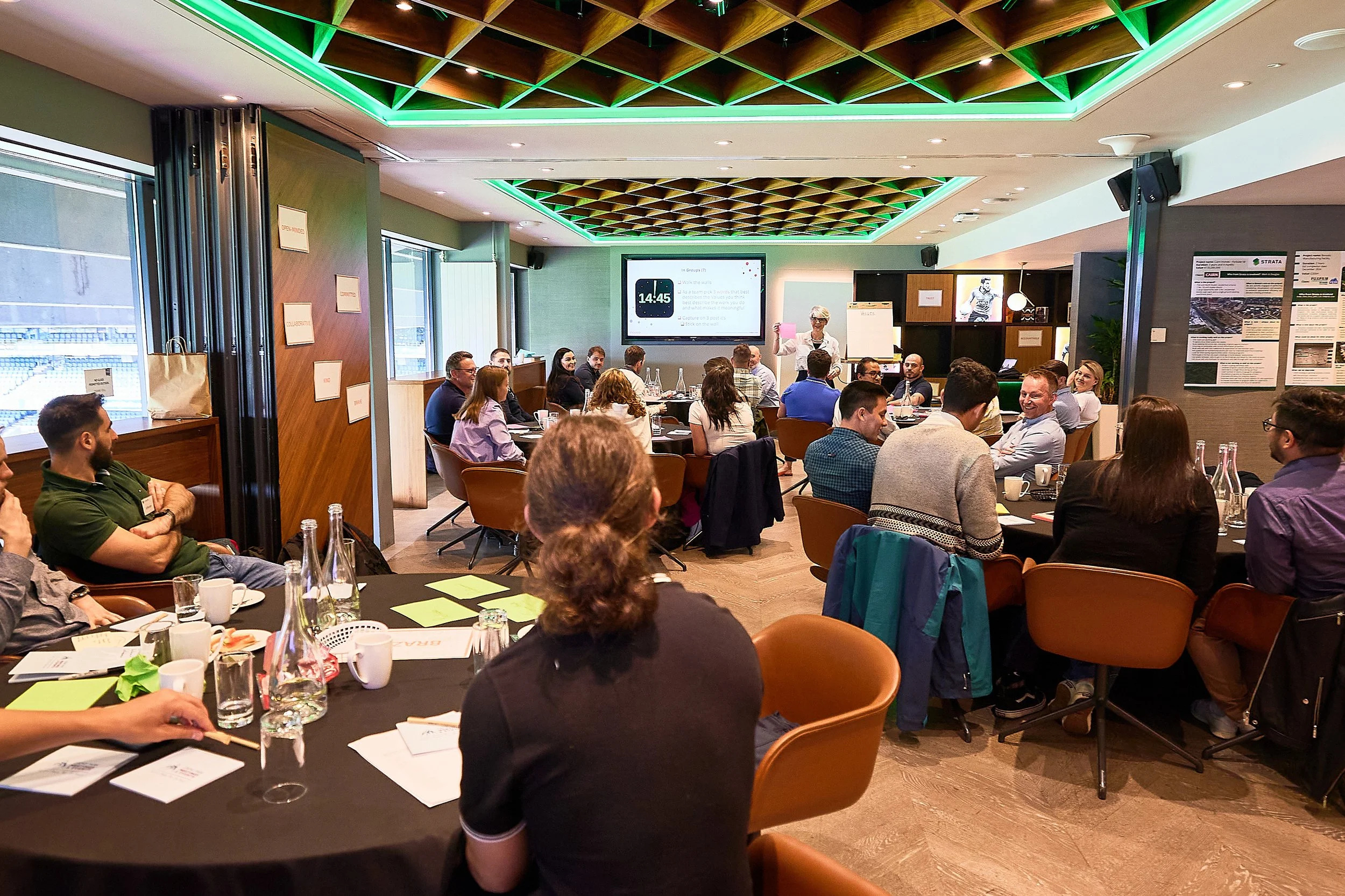 A group of profesional people attending a conference or meeting in a well lit room with a modern ceiling design, seated around tables, listening to a presenter standing at the front near a large screen.