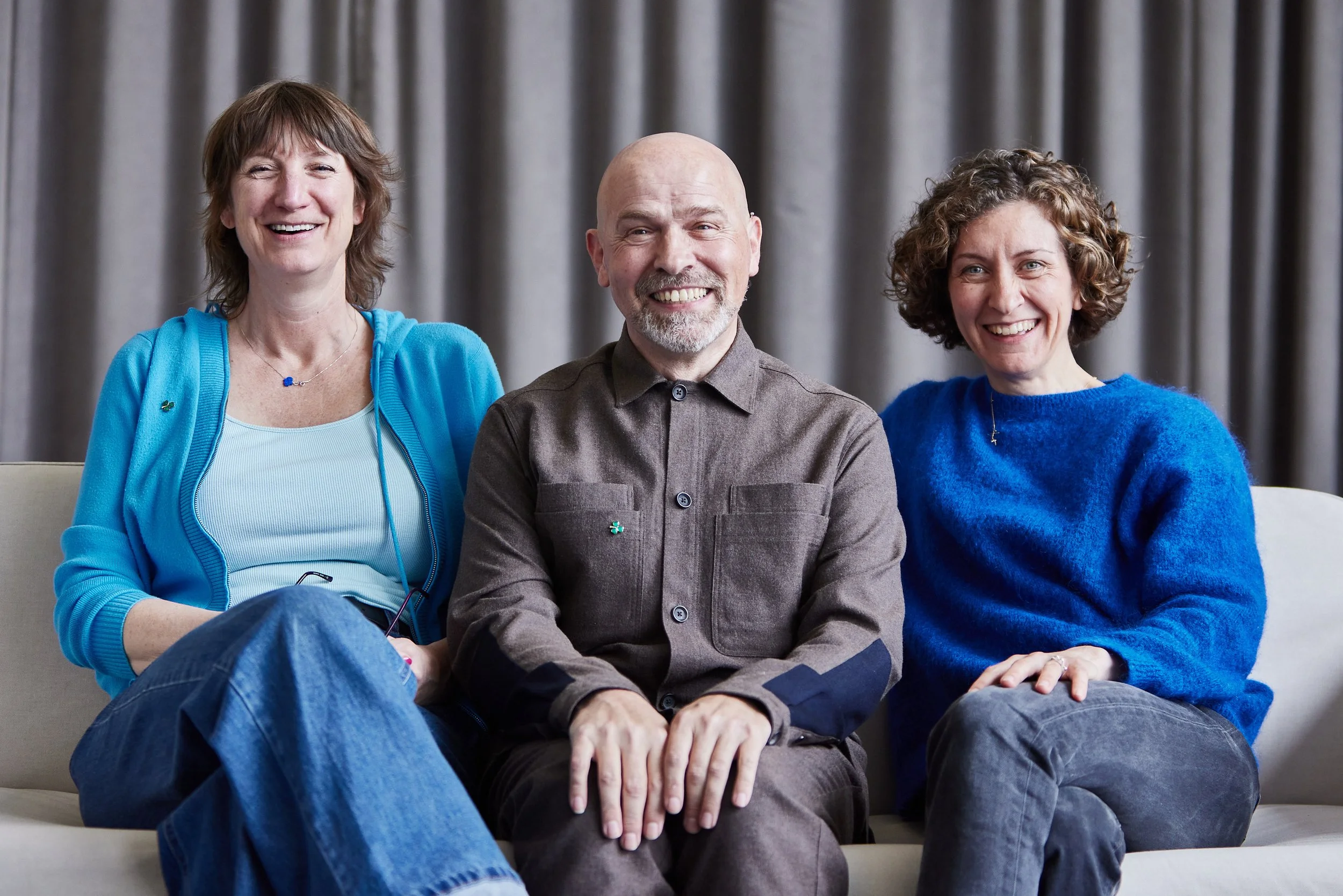 Three smiling adults sitting on a beige sofa in front of gray curtains. The person on the left is a woman with short brown hair, wearing a blue cardigan over a white top. The person in the middle is a man with a bald head and gray beard, wearing a br