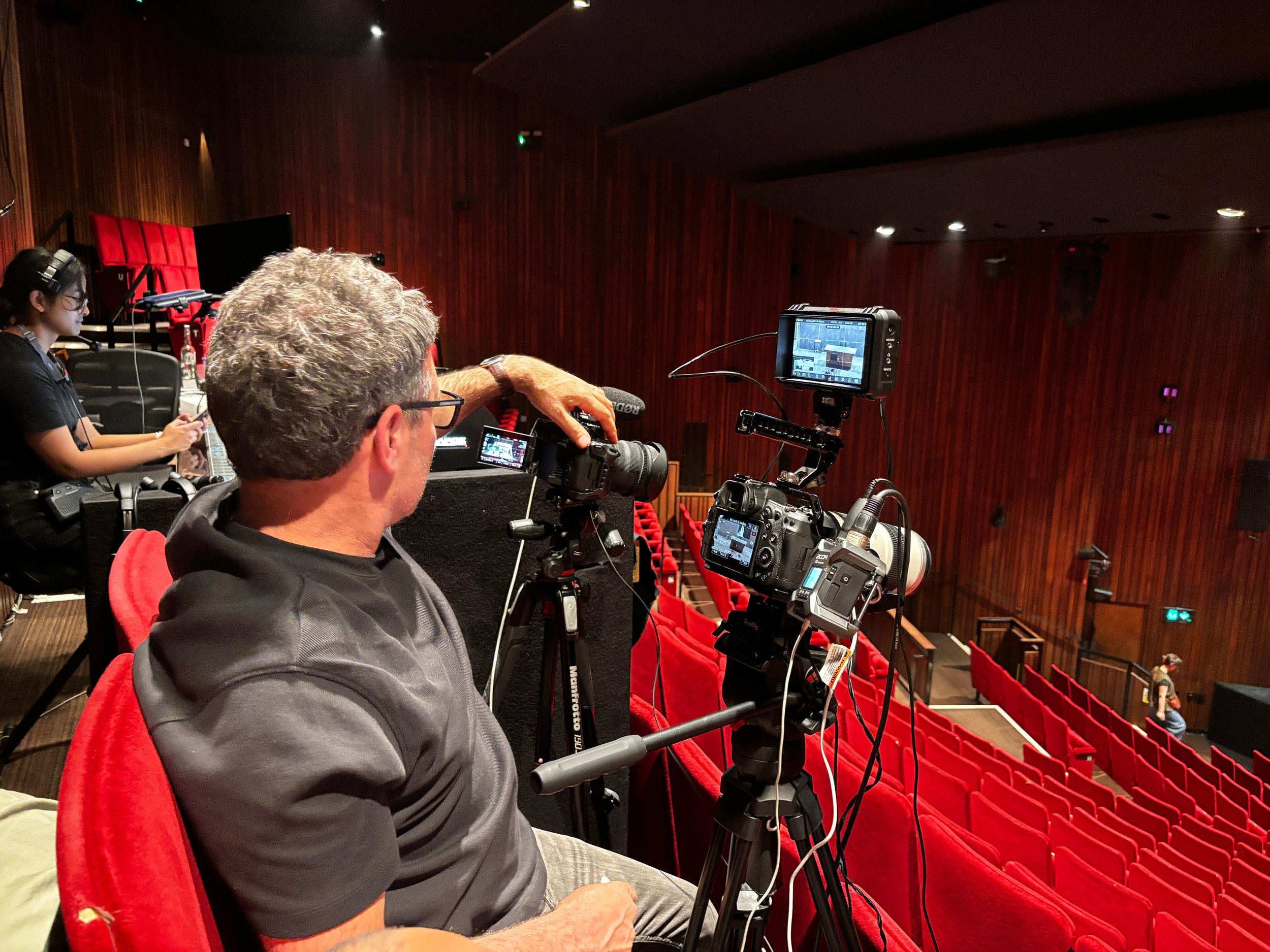 A man filming in an auditorium with red chairs and wood-paneled walls, accompanied by a woman working on a laptop in the background.