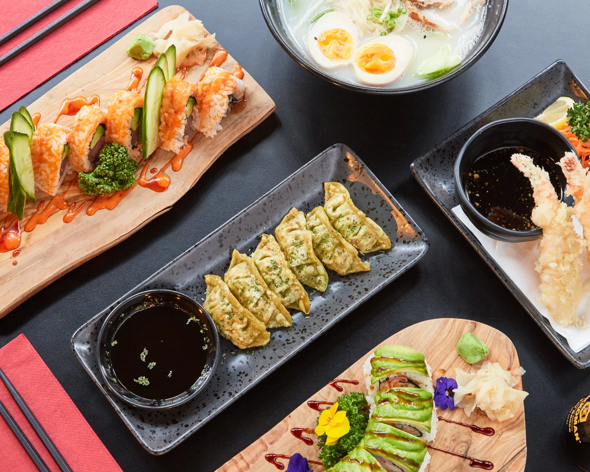 Advertising commercial professional photo of a variety of Japanese sushi dishes, including a sushi roll with avocado, a plate of gyoza dumplings with dipping sauce, tempura shrimp with dipping sauce, and a bowl of ramen on a black table.