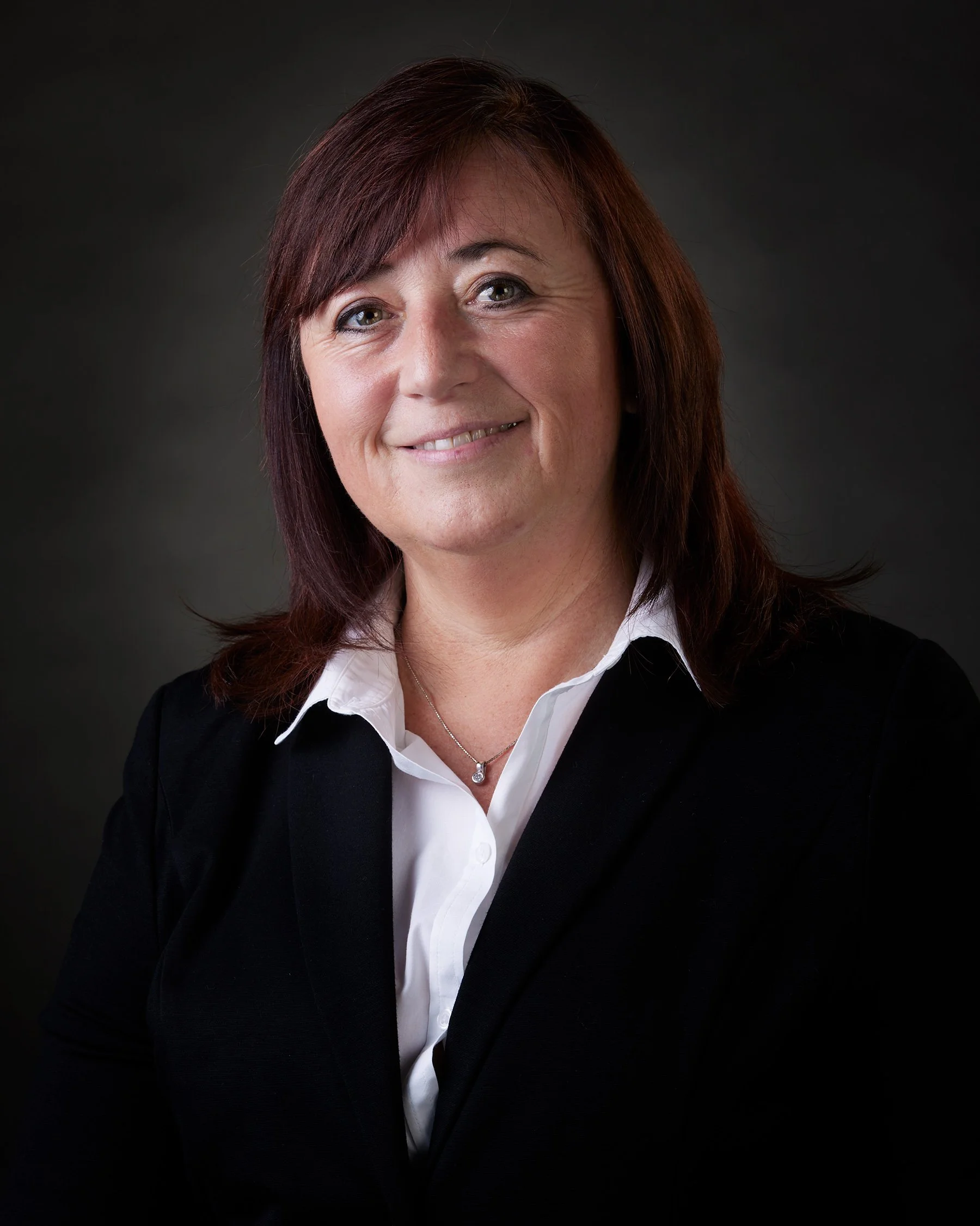 A professional headshot of a professional woman with shoulder-length red hair, wearing a black blazer over a white collared shirt, smiling against a dark background in a photo studio in a high quality lighted photo.