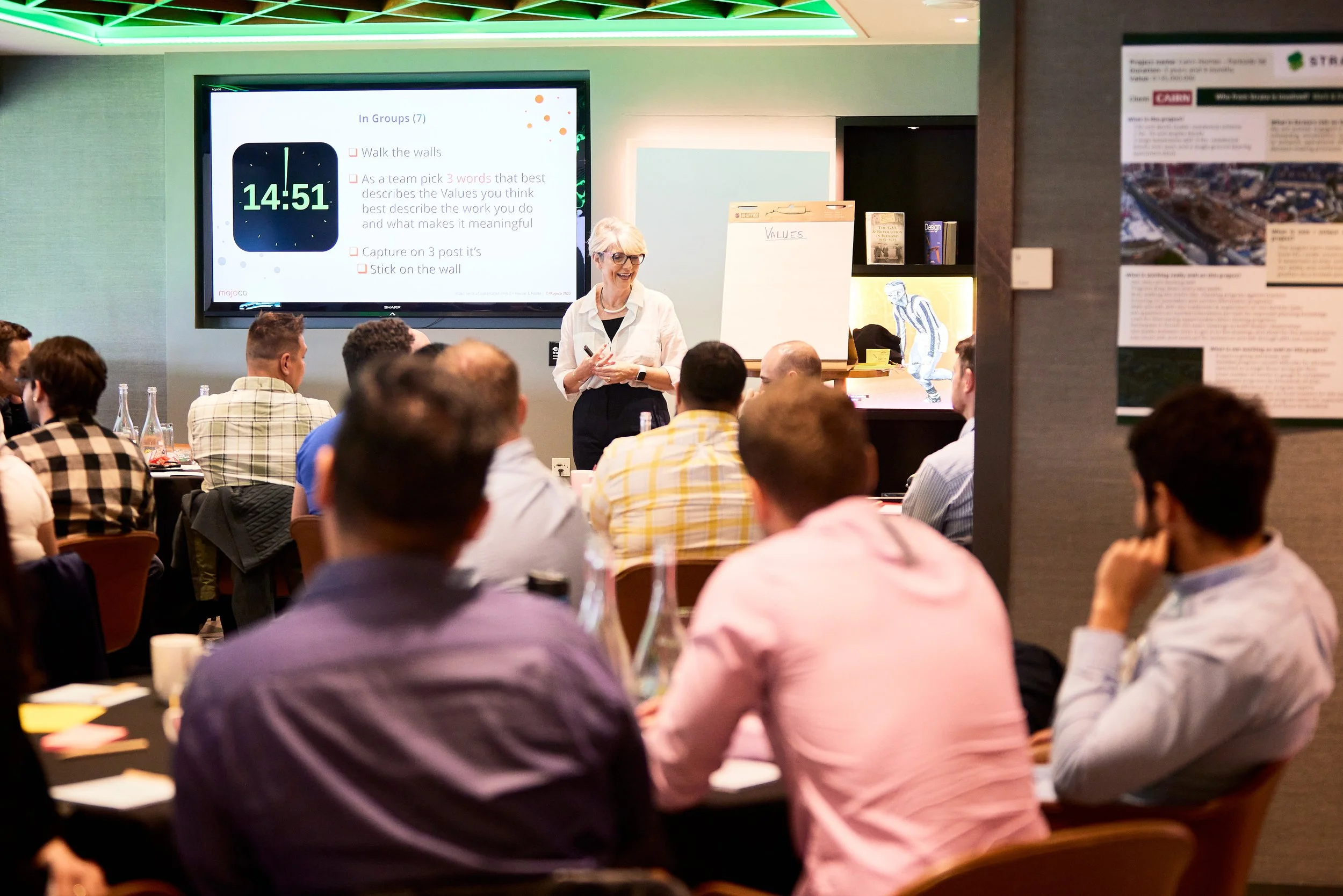 A woman giving a business presentation to a group of people seated at tables in a conference room. There is a large screen behind her displaying a slide with text and a clock showing 14:51. The audience appears engaged, with some taking notes.