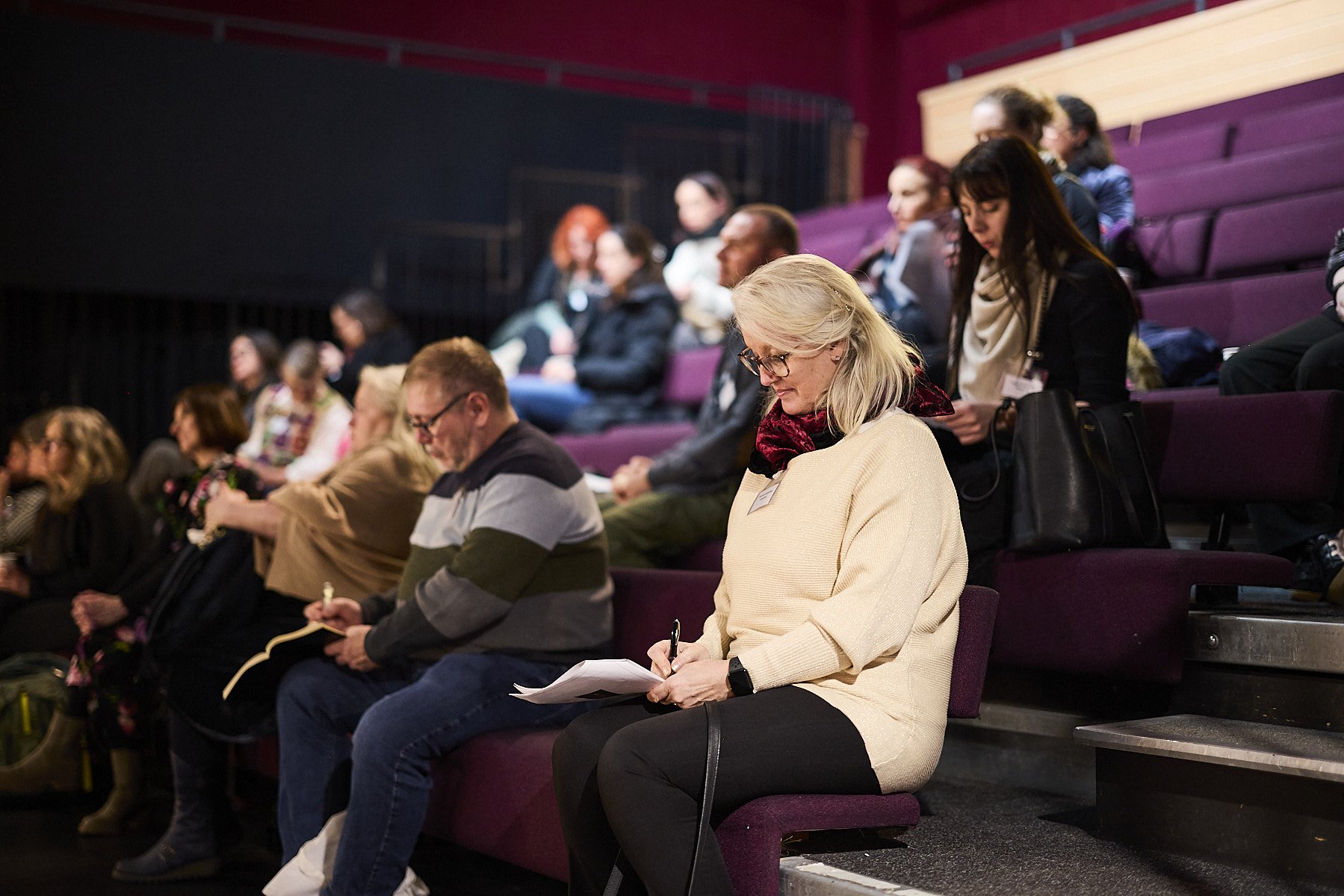 A group of people seated in purple theater seats attending an event, some are taking notes, in a darkened auditorium.