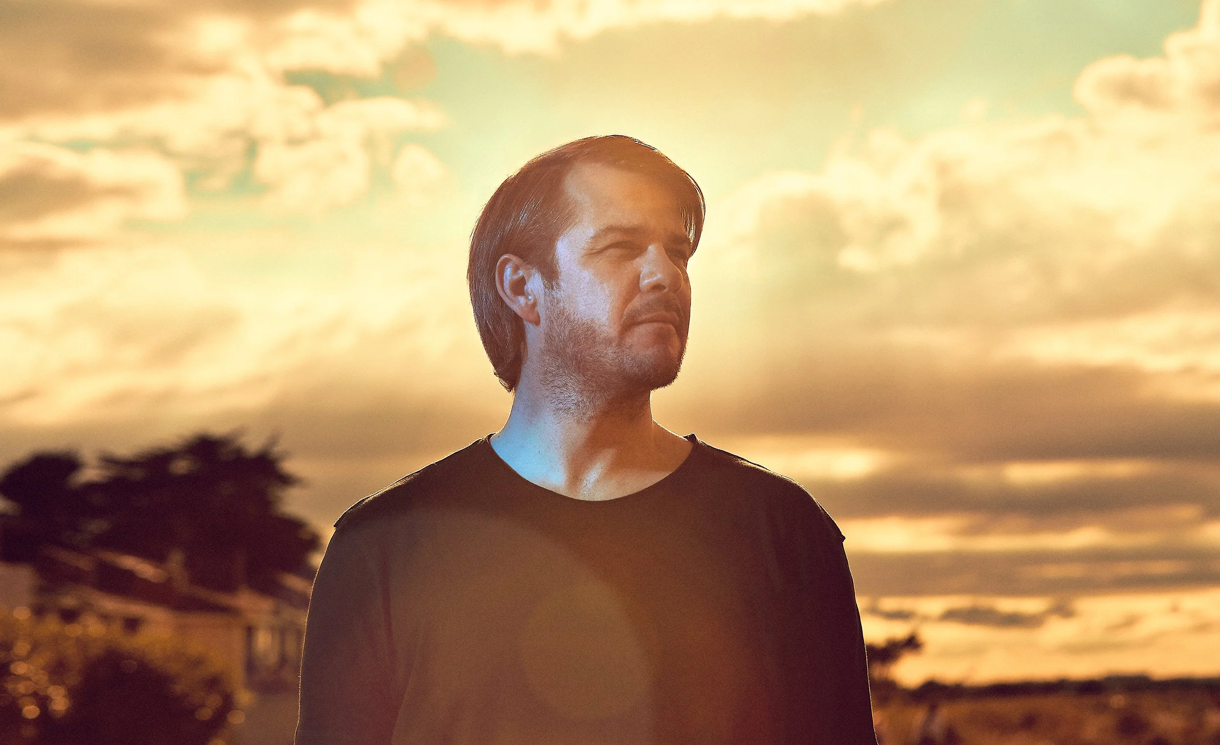 High quality advertising portrait of a man with dark hair and a beard stands outdoors during sunset or sunrise, with an orange sky and clouds in the background with professional photography lighting.