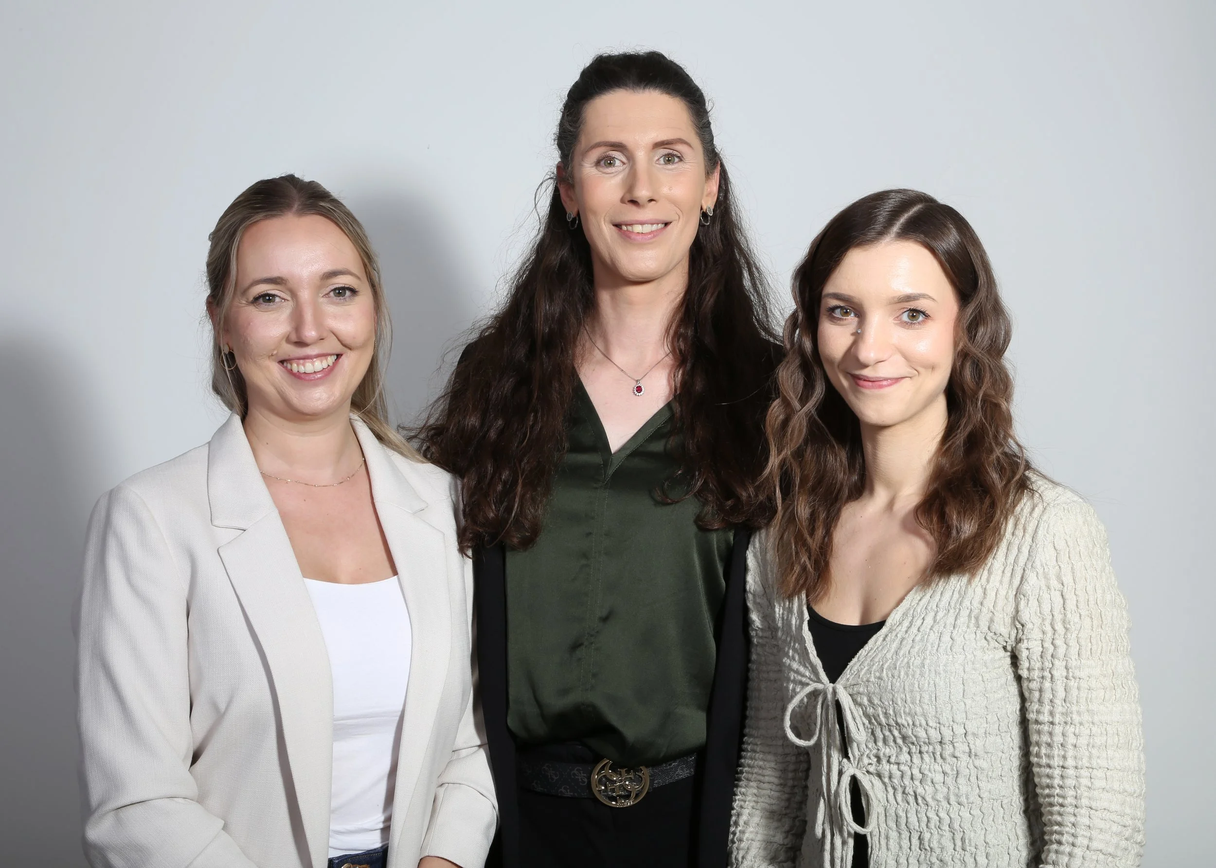 Drei Frauen stehen vor einem hellen Hintergrund und lächeln in die Kamera.