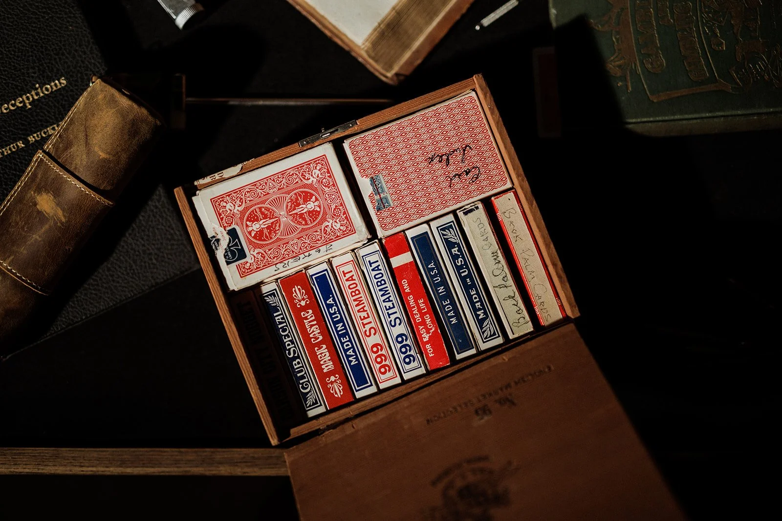 A wooden box containing several packs of vintage-style playing cards with red, blue, and white designs, including some with the logos and labeling of '99' and 'U.S.A.' sitting on a dark surface.