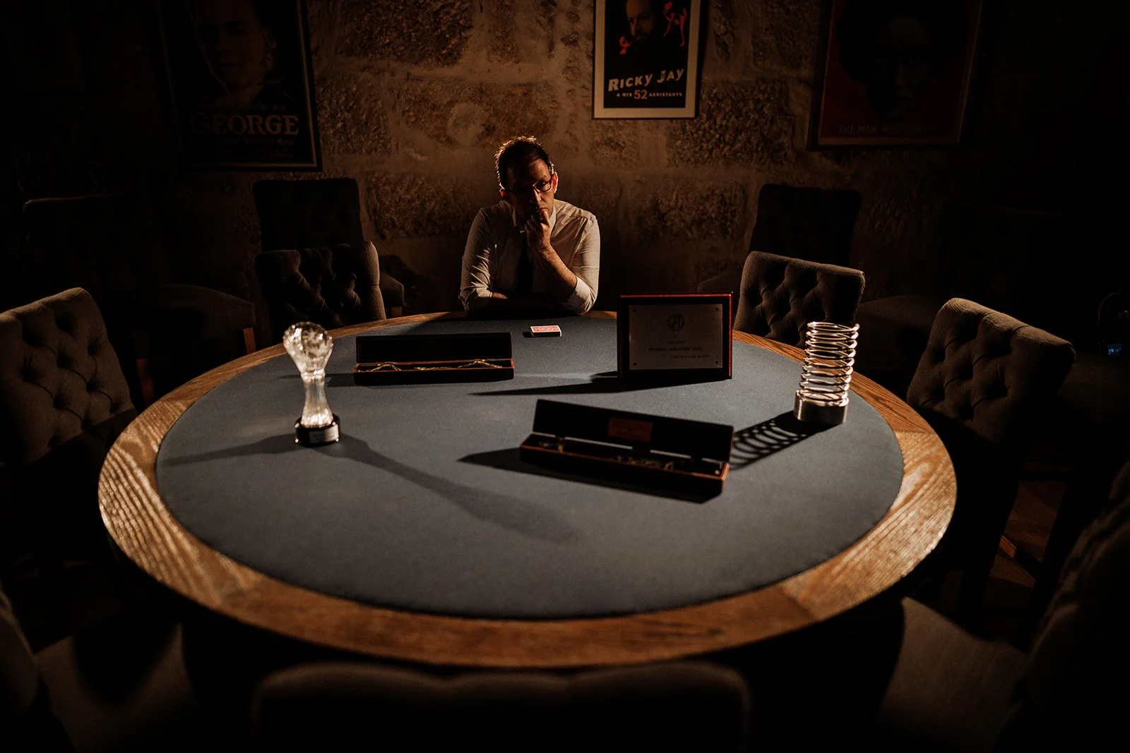 Helder Guimarães sitting alone at a round poker table in a dimly lit room, with his trophies, framed posters on the wall, and several chairs around the table.