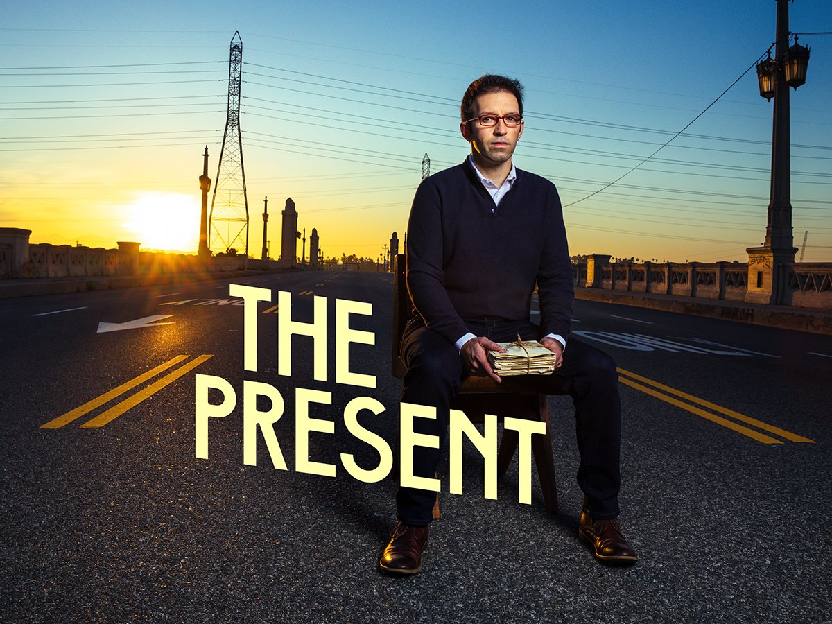 Helder Guimarães sitting on a chair in the middle of a road at sunset, holding a stack of money, with the text "The Present" overlaid.