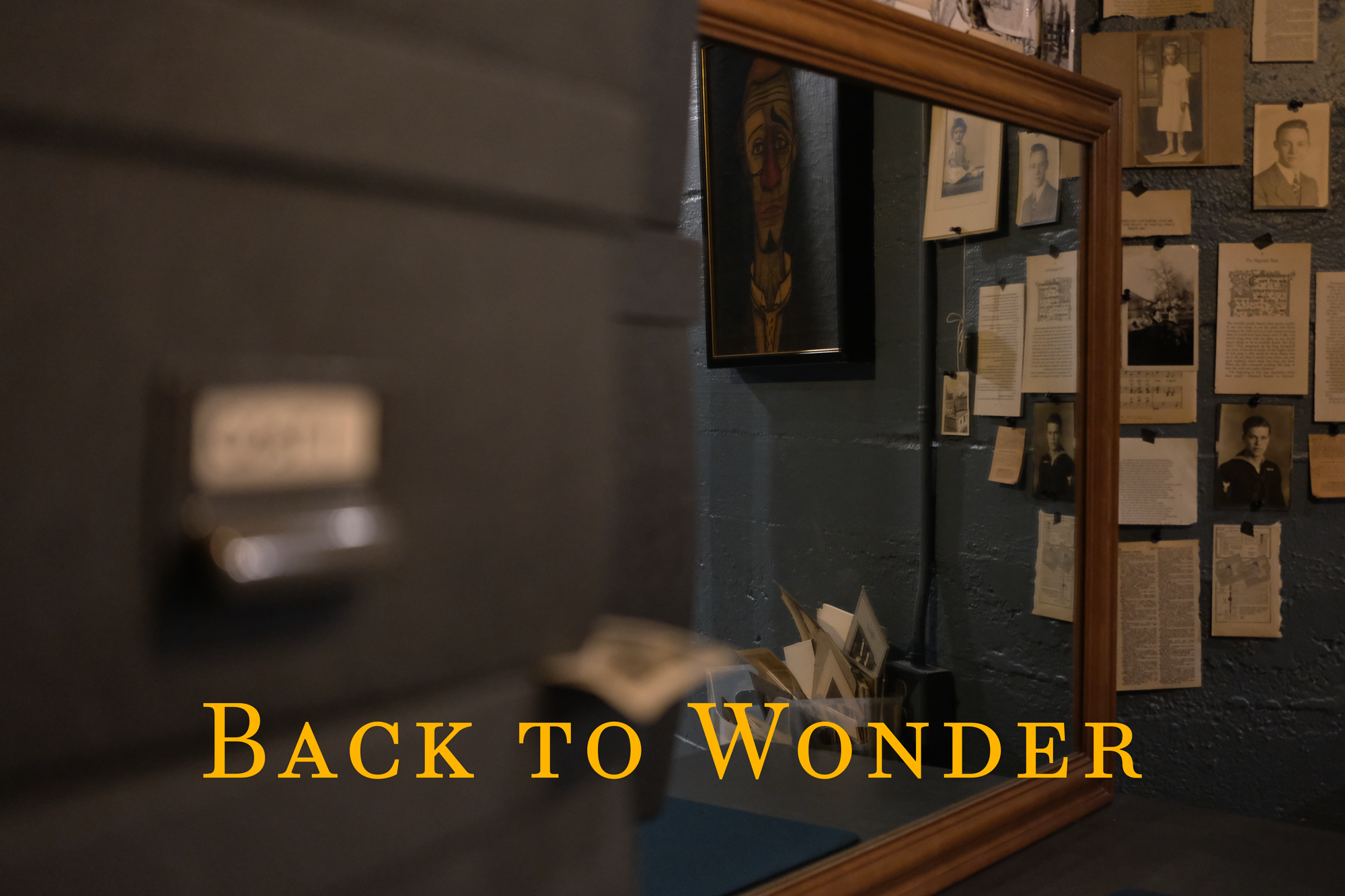 A museum exhibit with black and white photographs and historical documents on a wall, a framed painting of a clown's face, and a partial view of the text 'Back to Wonder' in bold orange letters at the bottom.