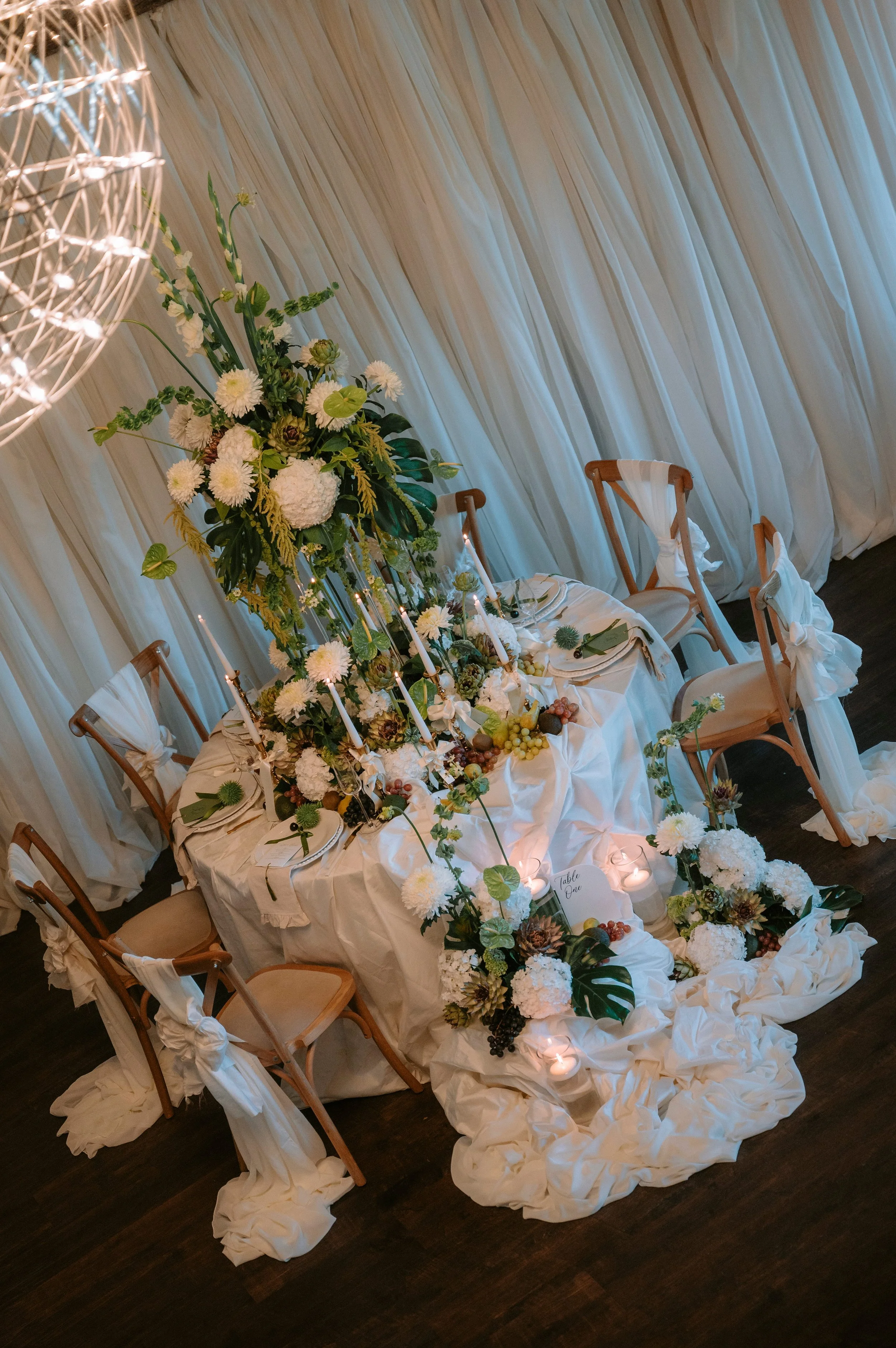 Decorated wedding reception table with white floral centerpiece, candles, grapes, and white draped fabric backdrop