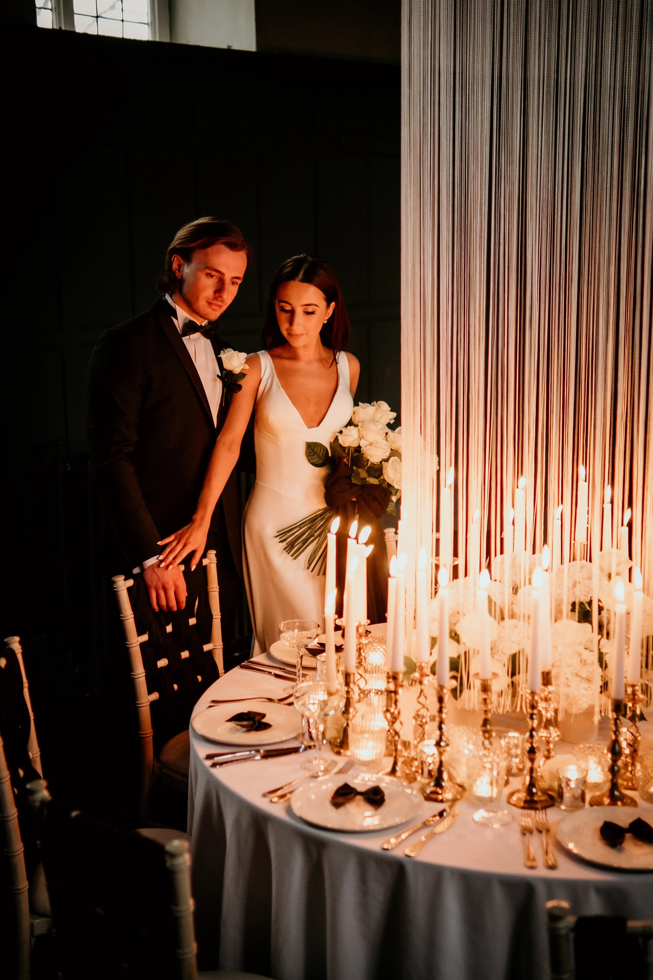 A bride and groom standing close together, looking at a table decorated with white candles, flowers, and elegant tableware, during a wedding reception. East Yorkshire based wedding florals, handcrafted wedding flowers.