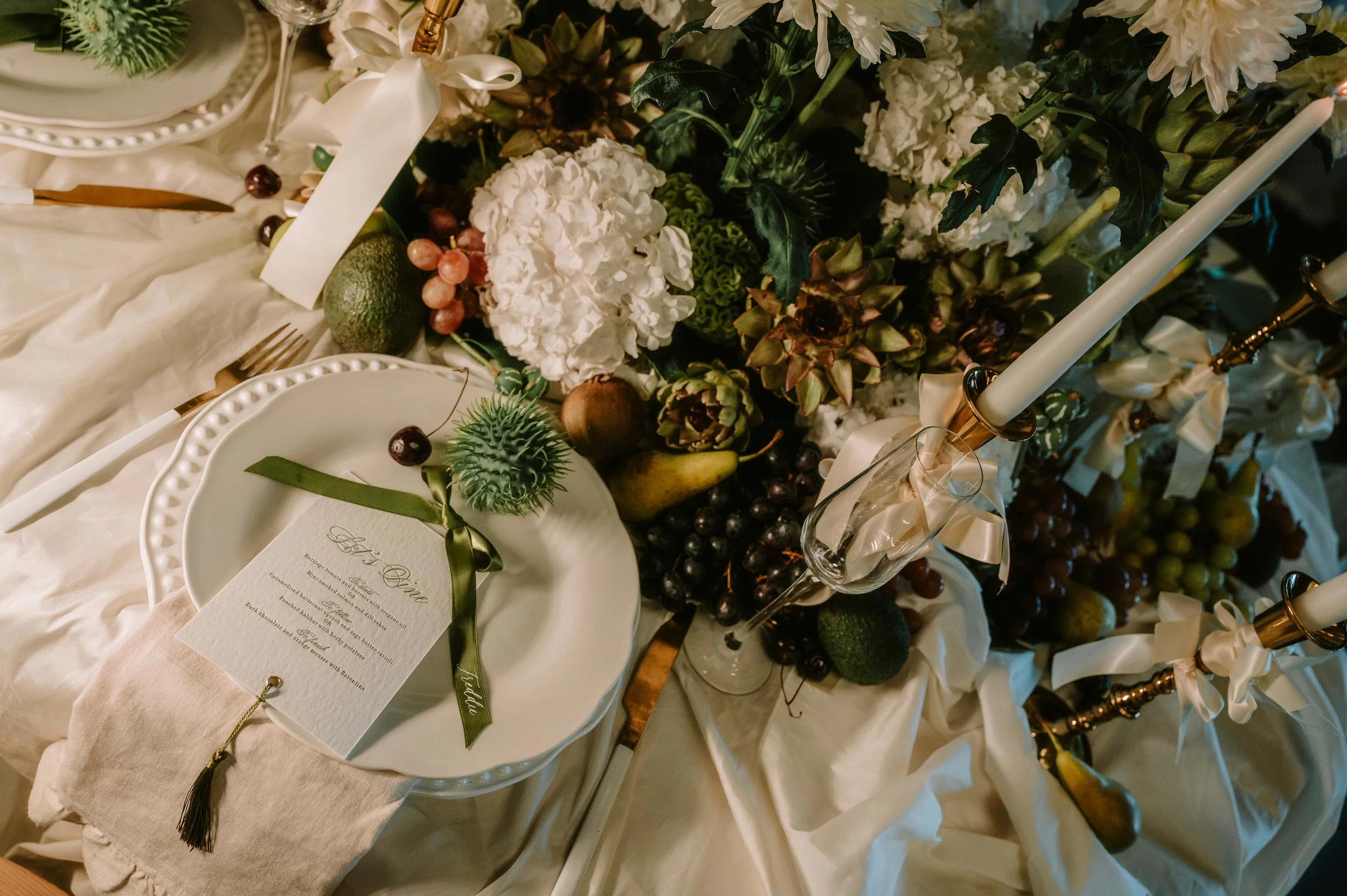 Elegant table setting with white plates, gold utensils, a menu card tied with a green ribbon, and a lush centerpiece of white hydrangeas, grapes, and other flowers, accented with tall white candles in gold holders.