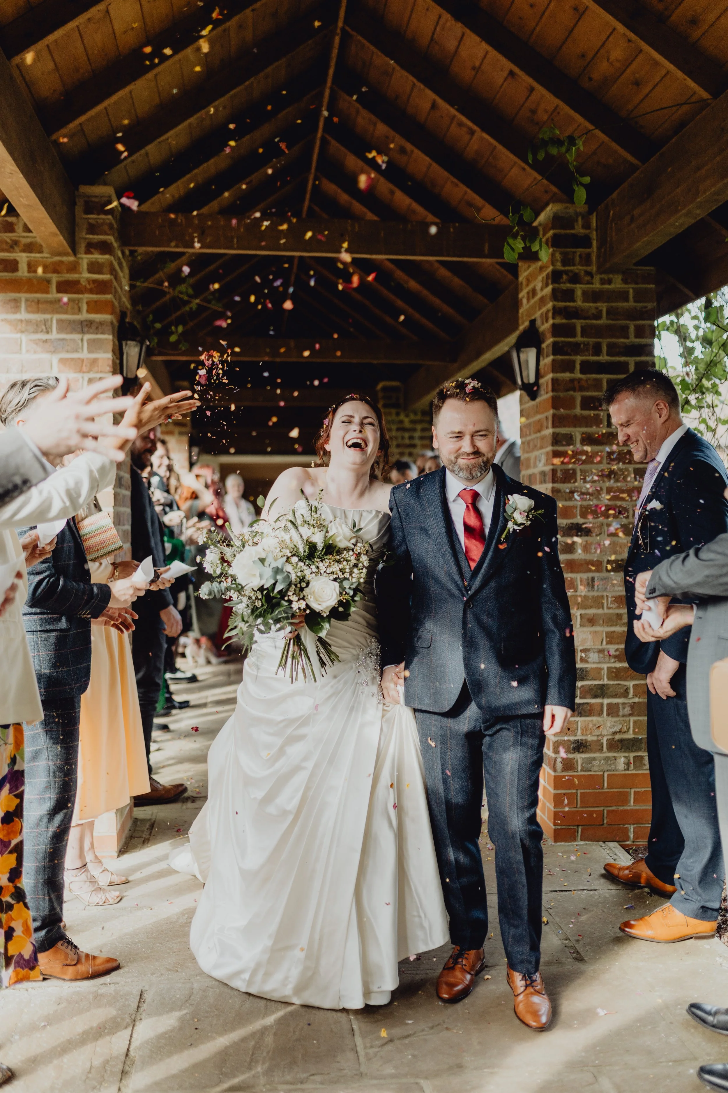 East Yorkshire based wedding florals, handcrafted wedding flowers. Bride and groom walking through confetti under a wooden roof, smiling, surrounded by guests. Vicky Johnston