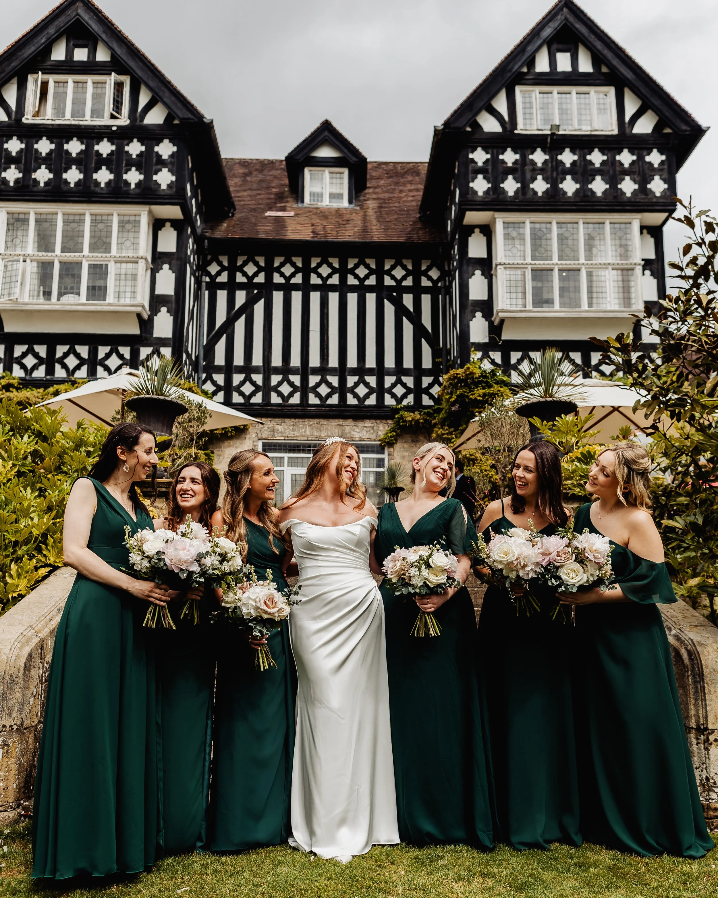 A bride in a white wedding dress surrounded by six bridesmaids in green dresses, all holding bouquets of flowers, standing in front of a large timber-framed house and garden with umbrellas and plants. East Yorkshire based wedding florals, handcrafted