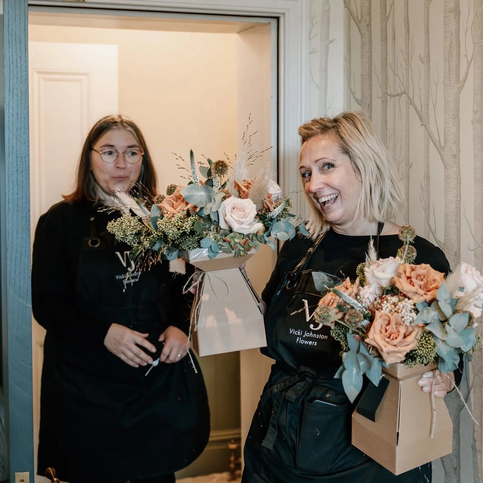 Two women smiling and holding flower arrangements in boxes, inside a room with patterned wallpaper. East Yorkshire based wedding florals, handcrafted wedding flowers.