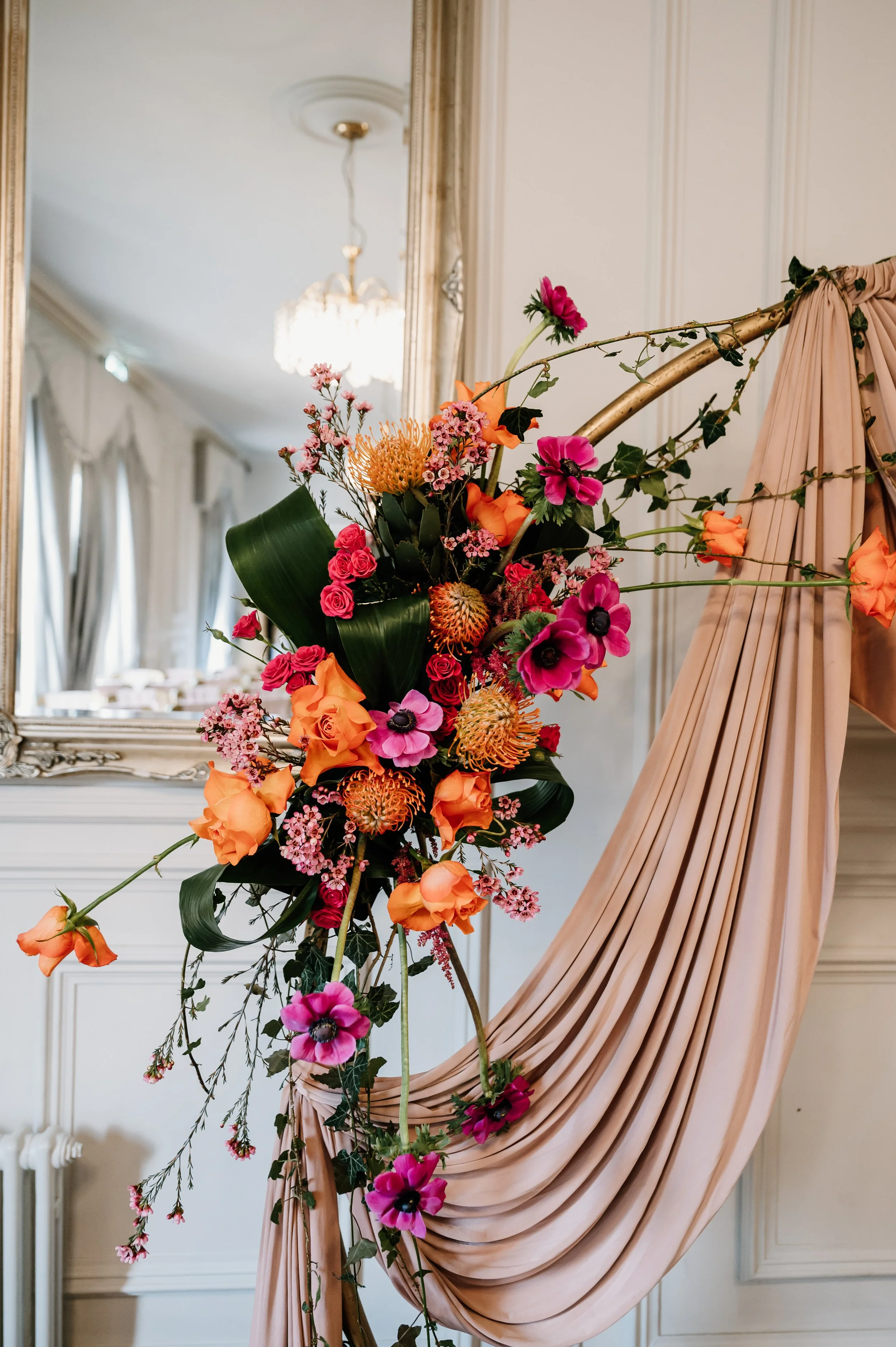 Elegant staircase decorated with a lush floral arrangement featuring pink, orange, and red flowers, with a draped blush-colored fabric, all set in a sophisticated interior with ornate mirror and chandelier. East Yorkshire based wedding florals.