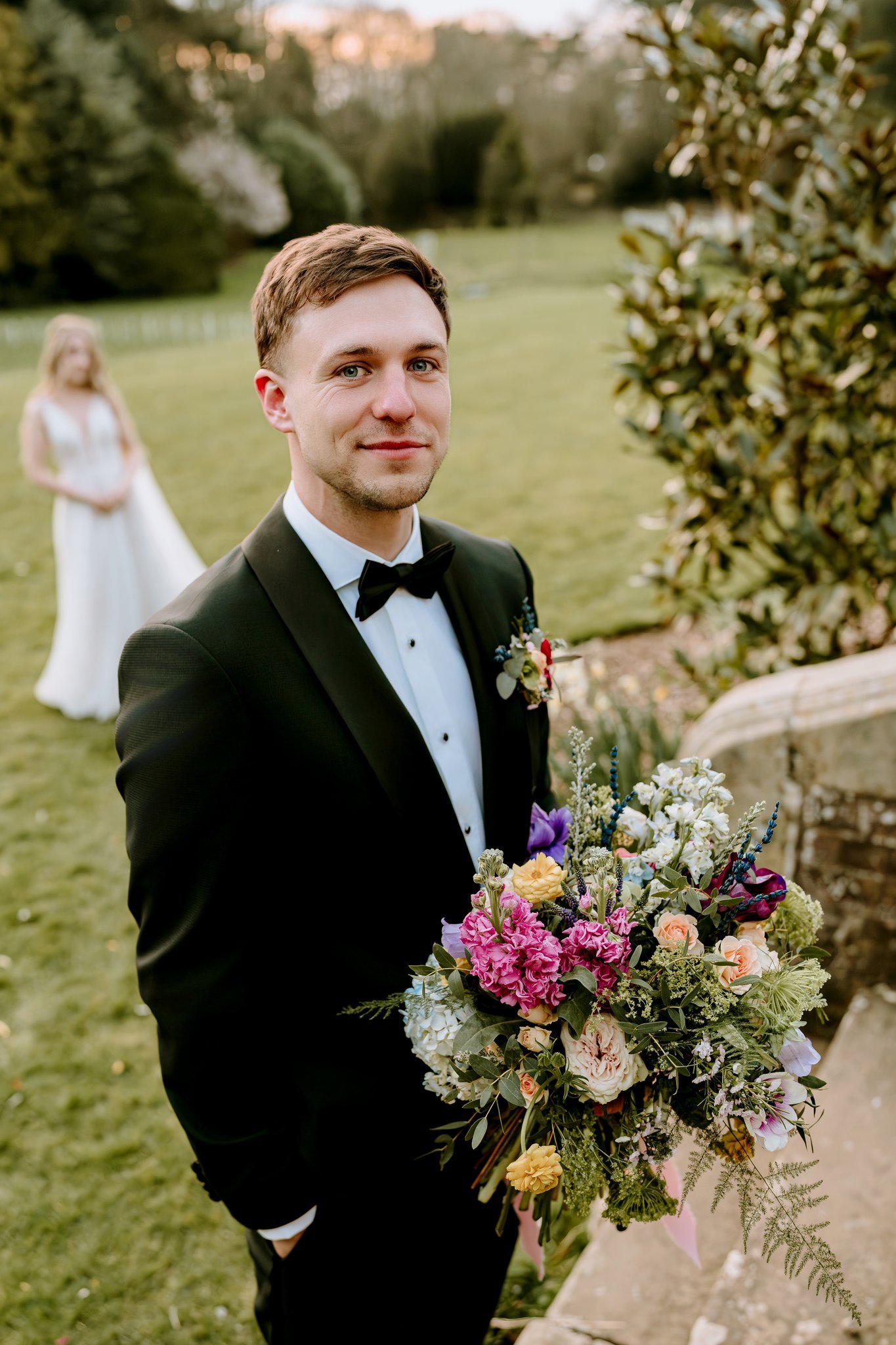 A groom in a black tuxedo holding a colorful bouquet of flowers, standing outdoors in a garden with a blurred bride in a white wedding dress in the background. East Yorkshire based wedding florals, handcrafted wedding flowers.