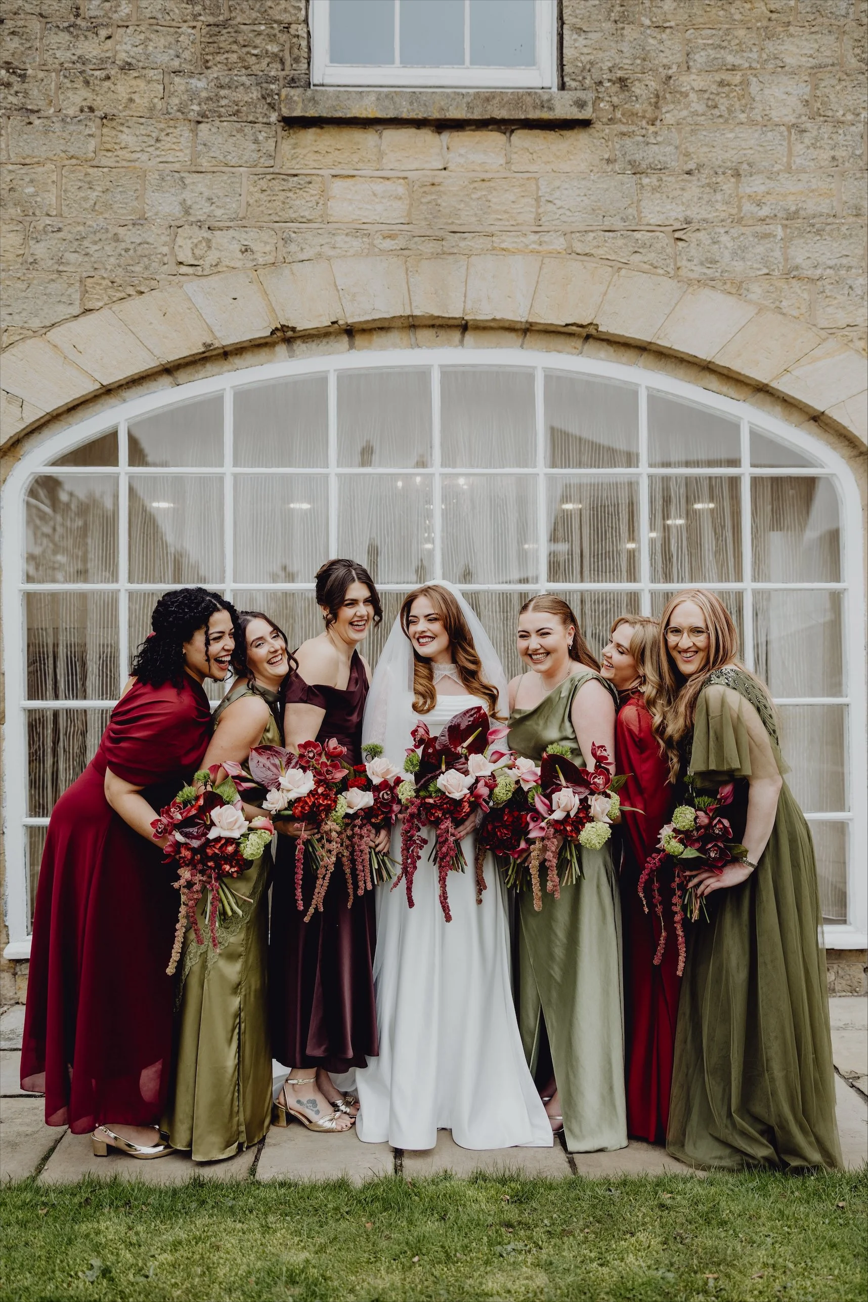 Group of seven women, including bride in white gown, smiling and holding bouquets, standing outside near a large window with a stone building in the background.