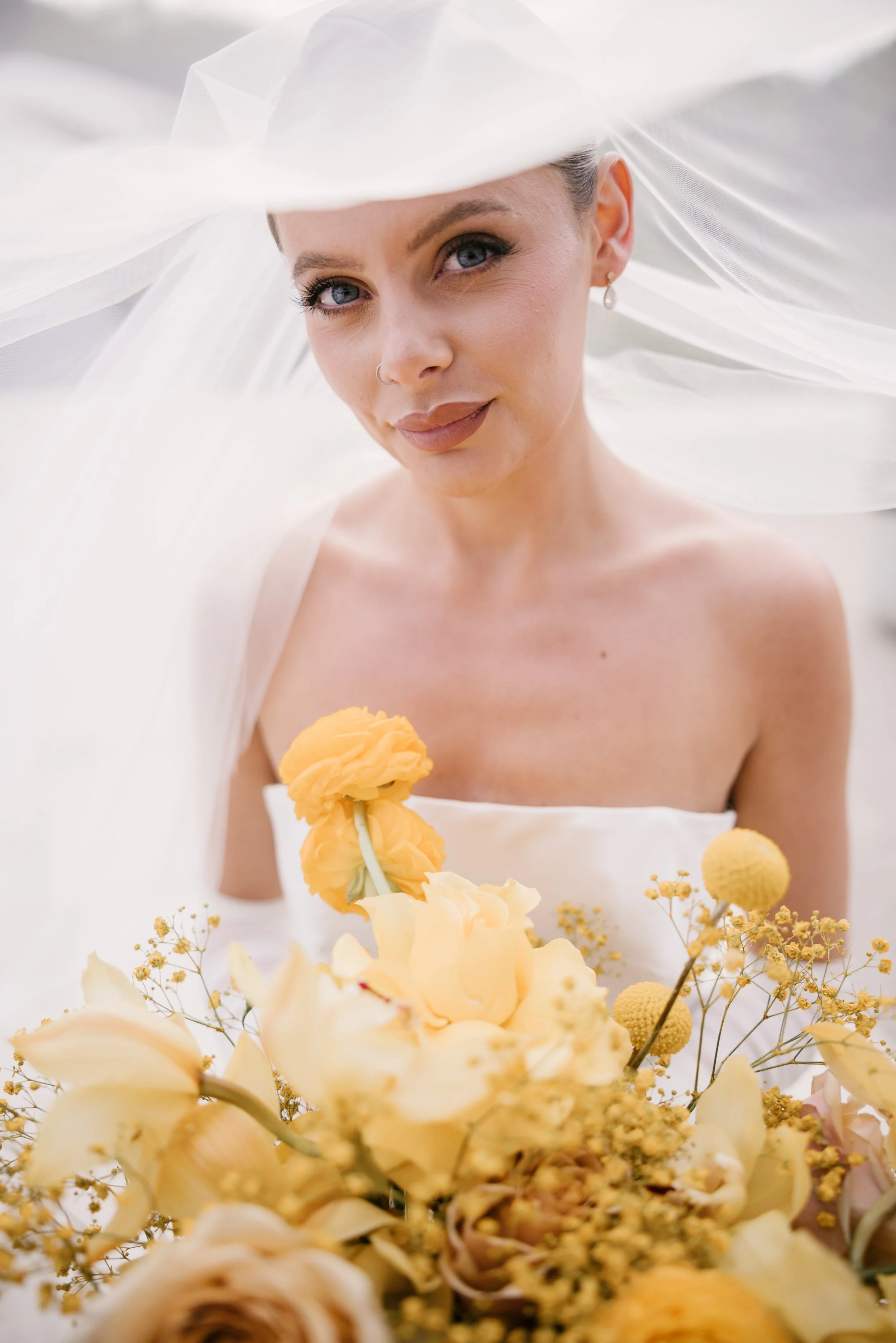 A bride with blue eyes and light makeup, wearing a white strapless dress and a large white veil, holding a bouquet of yellow and cream flowers, and smiling softly. East Yorkshire based wedding florals, handcrafted wedding flowers.