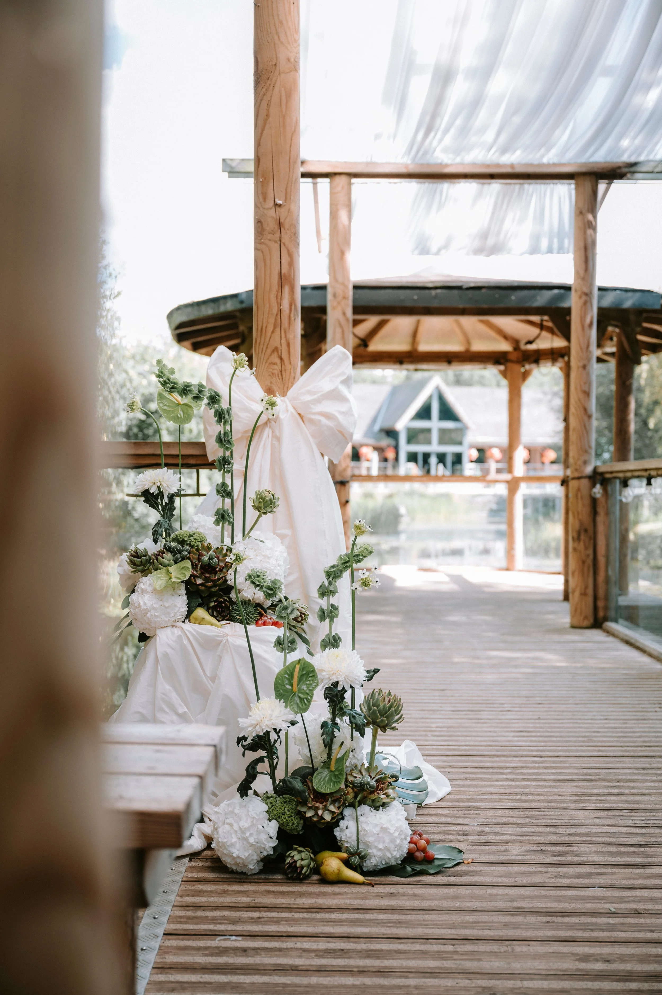 Decorative arrangement of white flowers, candles, fruits, and greenery on a draped table on a wooden walkway in an outdoor setting with a wooden gazebo and building in the background.