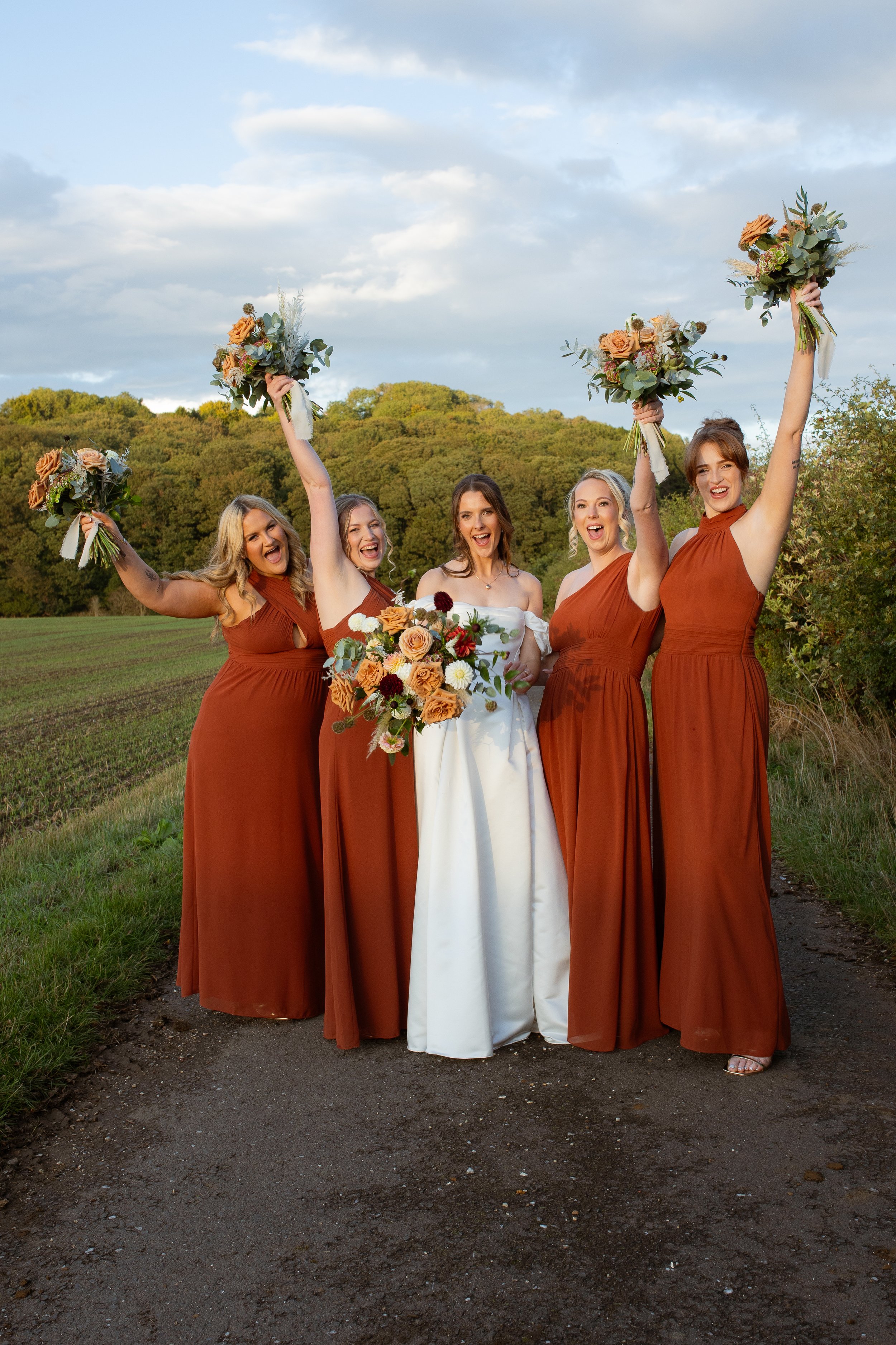 Group of five women celebrating outdoors, with three bridesmaids in rust-colored dresses holding bouquets and a bride in a white dress holding a bouquet, standing on a dirt path surrounded by fields and trees with a partly cloudy sky.