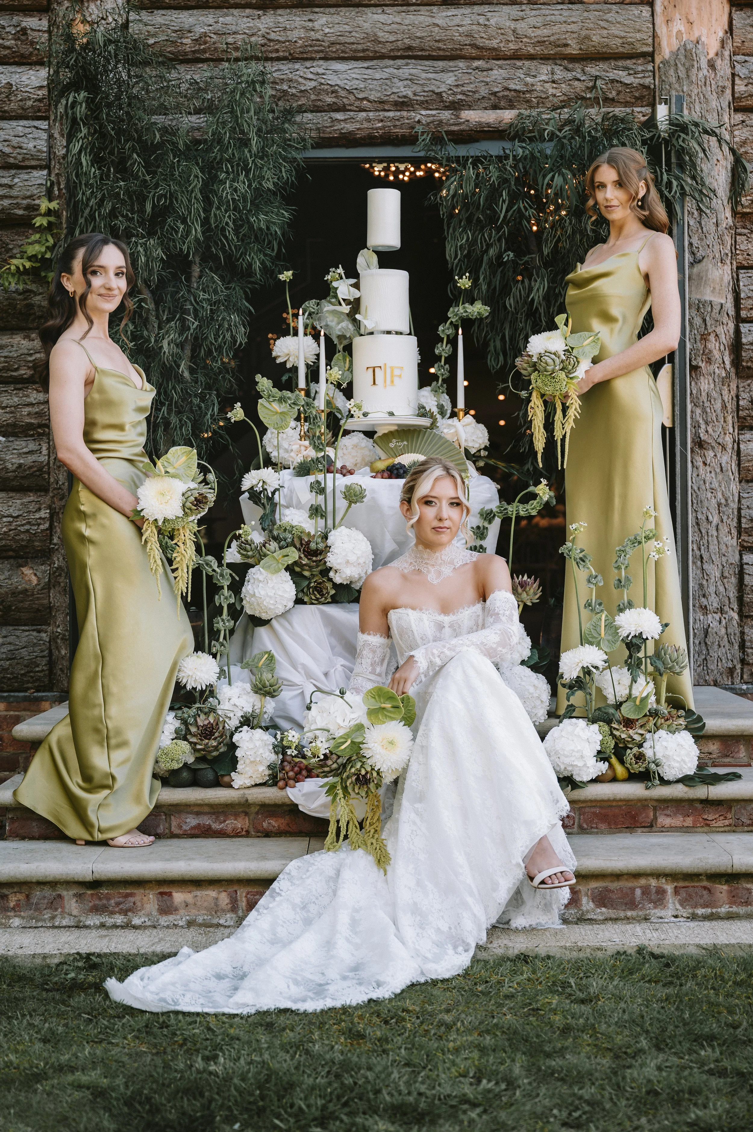 A bride in a white wedding dress sitting on the steps in front of a decorated table with white flowers and a tiered wedding cake, flanked by two bridesmaids in matching gold satin dresses holding bouquets, at an outdoor wedding. East Yorkshire floral