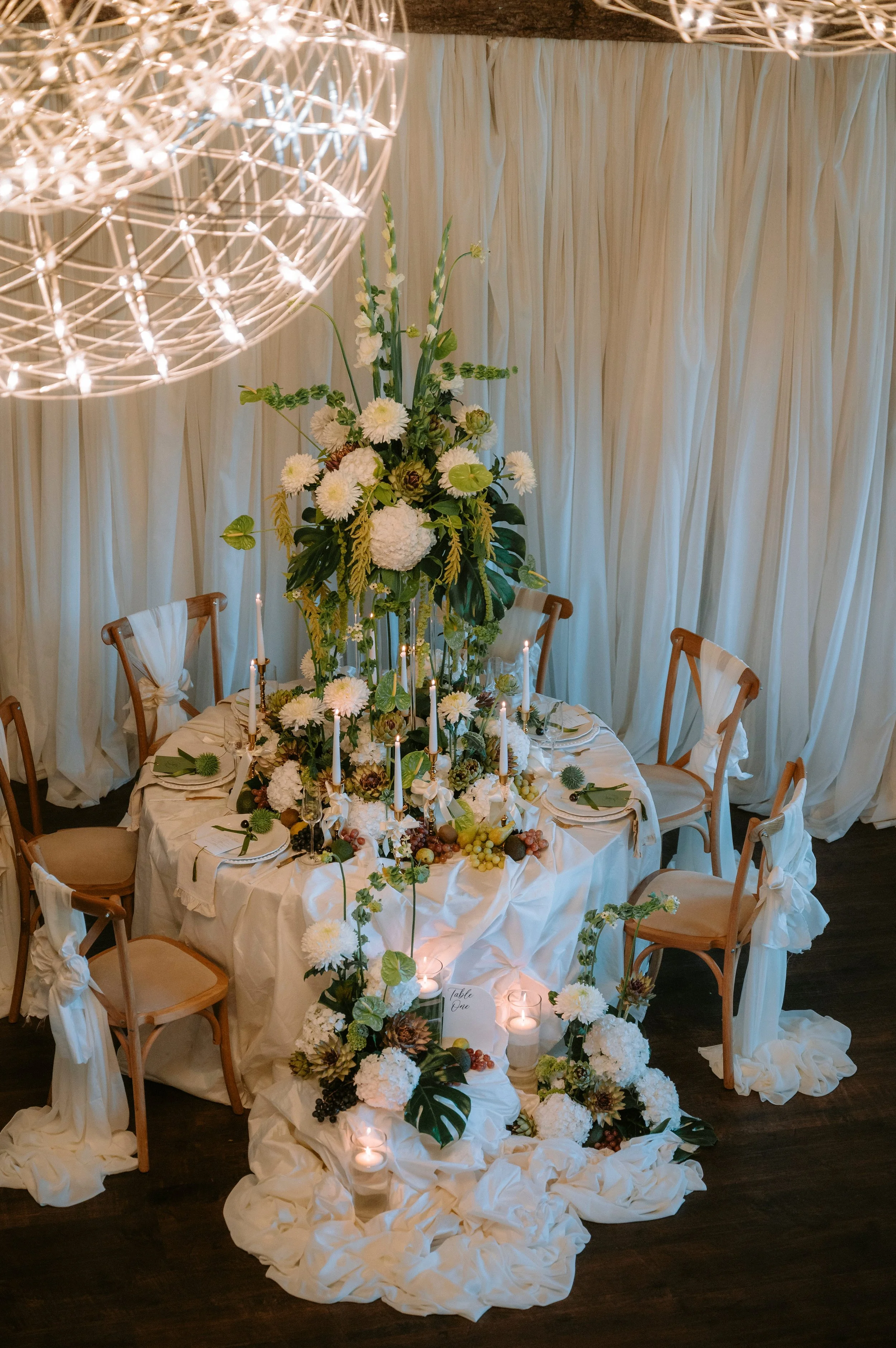Round dining table decorated with a tall floral centerpiece, surrounded by white curtain backdrop, candlelit accents, and beige chairs with white fabric tied to the backs, set for a formal event.