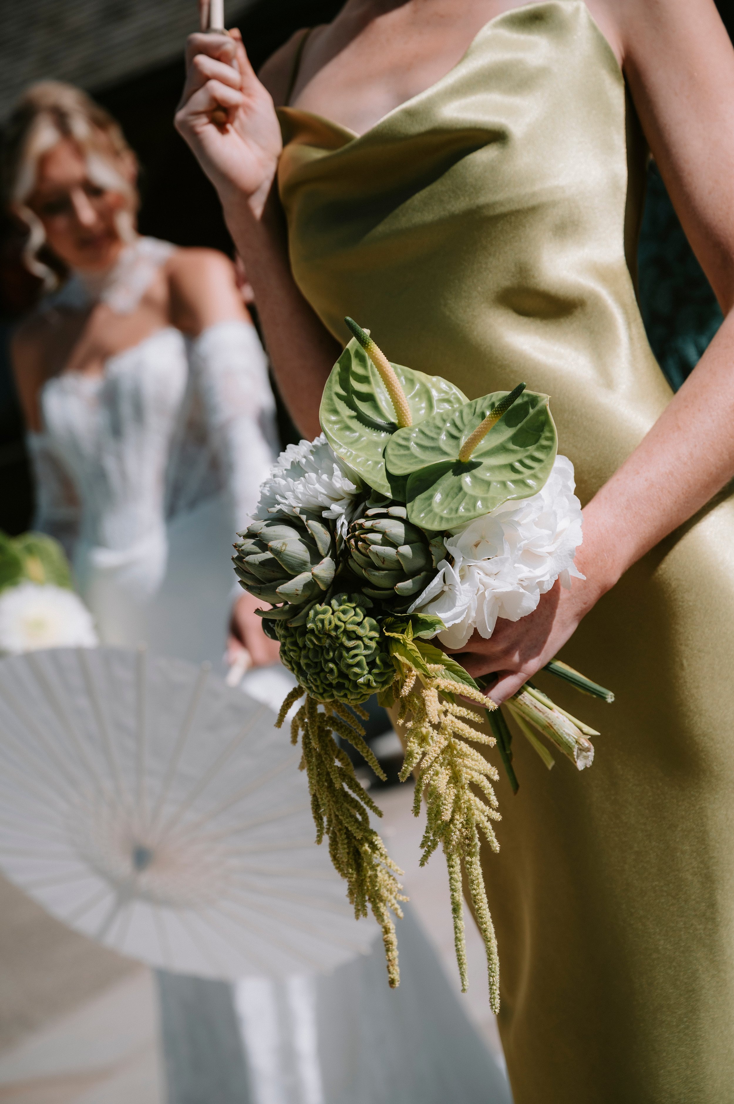 A woman in a gold satin dress holding a bouquet of various green and white flowers at a wedding or special event, with another woman in a white dress blurred in the background.