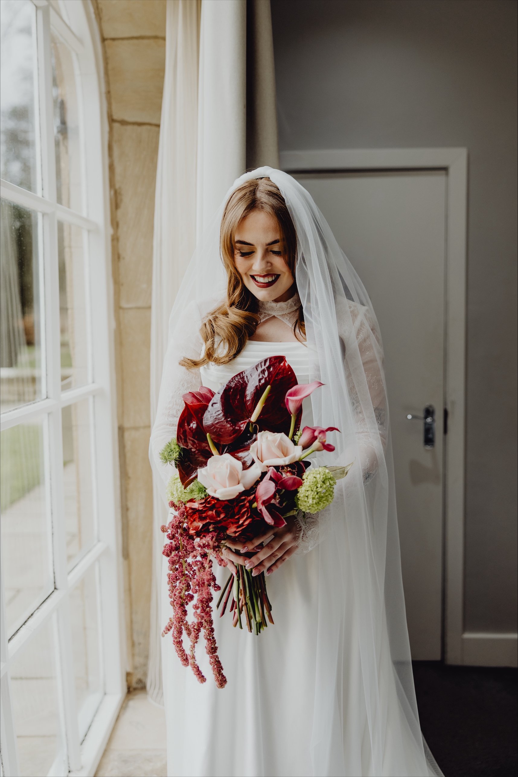 A bride in a white wedding dress and veil holding a bouquet of pink and red flowers, standing near a window with cream-colored curtains, smiling and looking down.