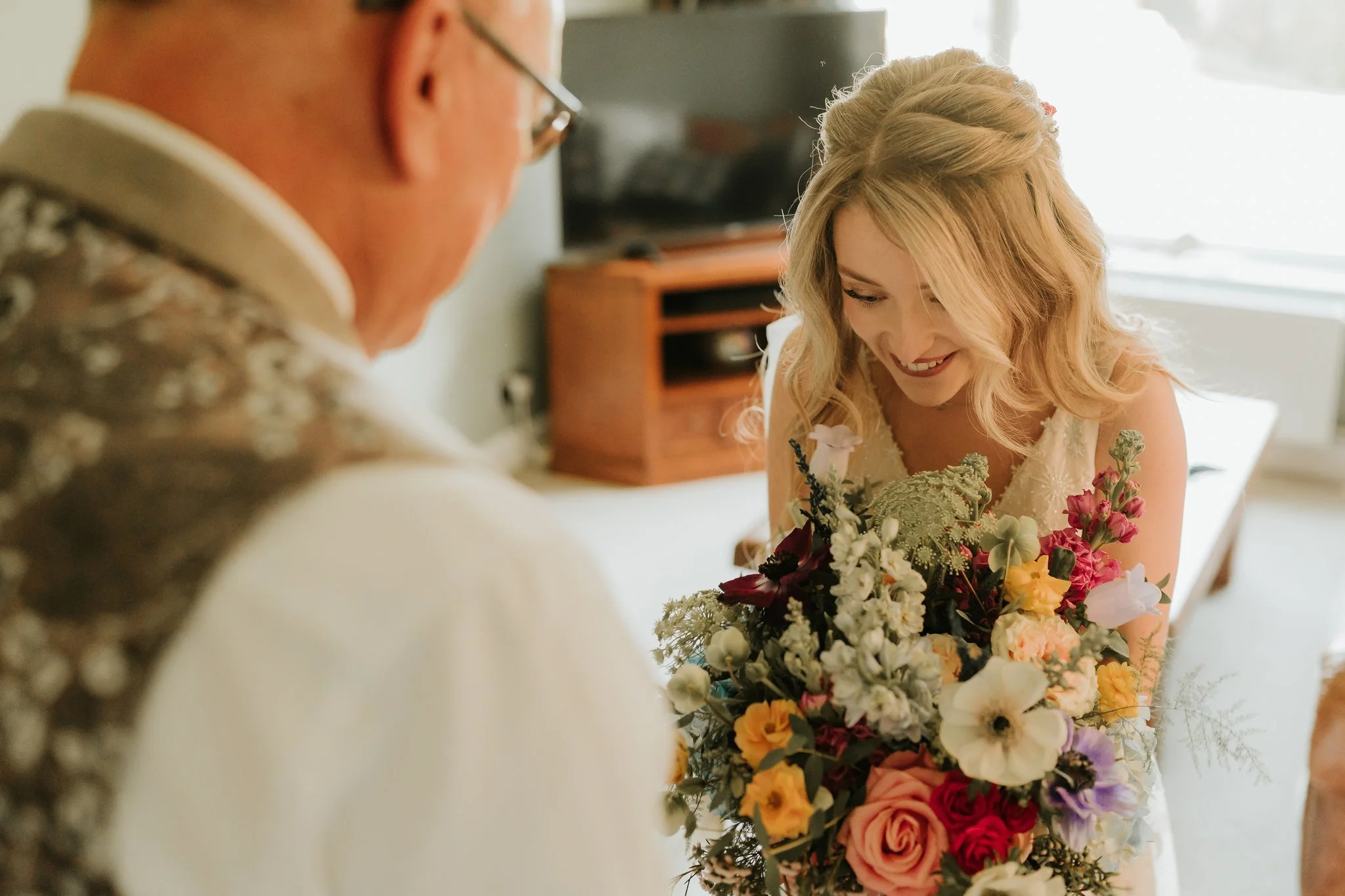 A woman in a white dress holding a large bouquet of colorful flowers, smiling as she looks down, while a man in glasses and a patterned vest looks on in a cozy indoor setting. East Yorkshire based wedding florals, handcrafted wedding flowers.